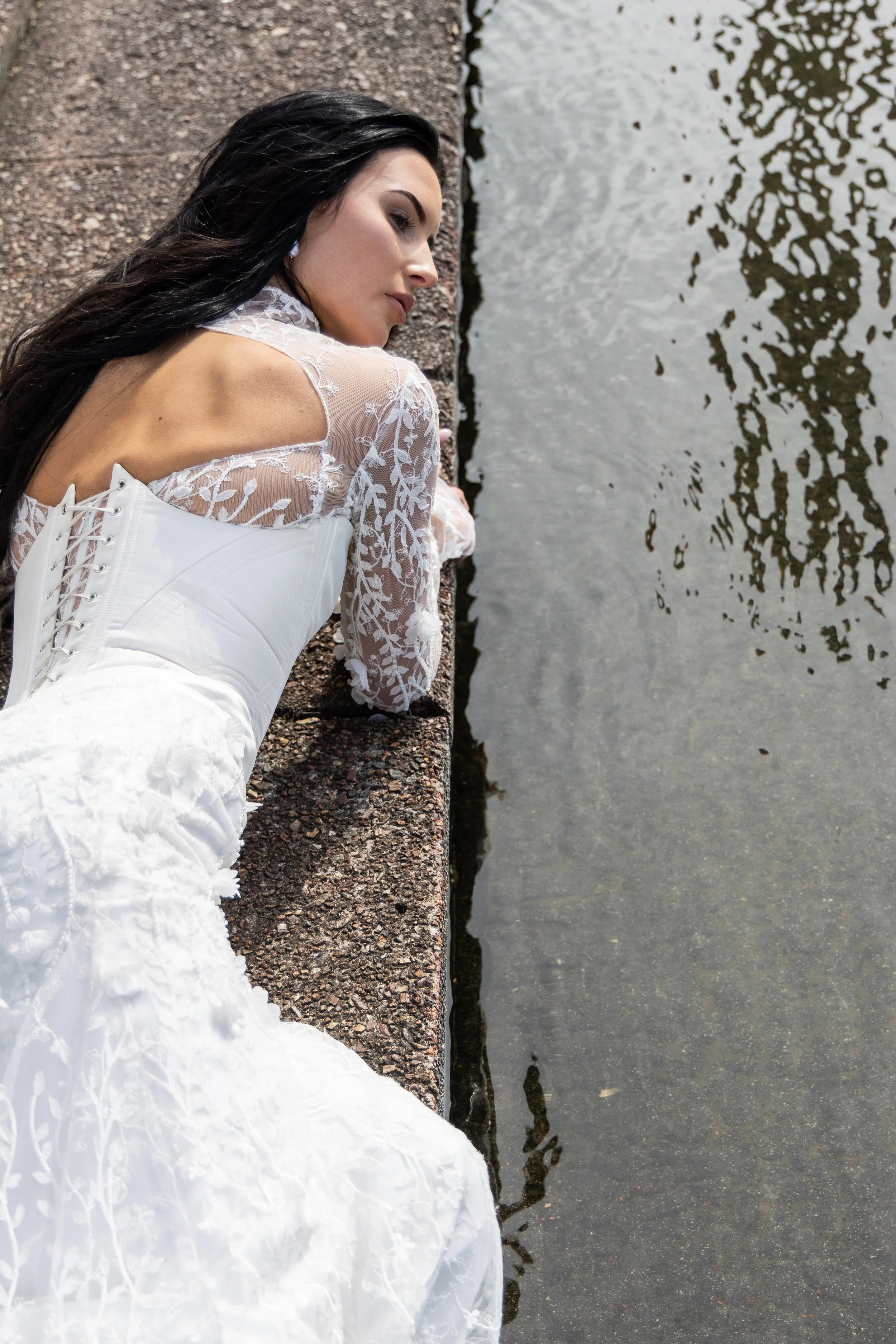 A woman with long dark hair is lying on her side on a concrete surface next to a body of water, wearing a white embroidered dress with lace-up details on the back.