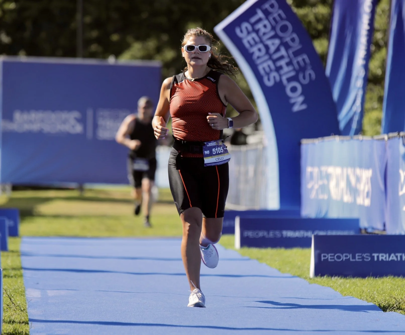 A woman running in a triathlon event on a blue track, wearing athletic gear and sunglasses, with two other runners behind her. Banners with the event name 'PEOPLE'S TRIATHLON' are visible.