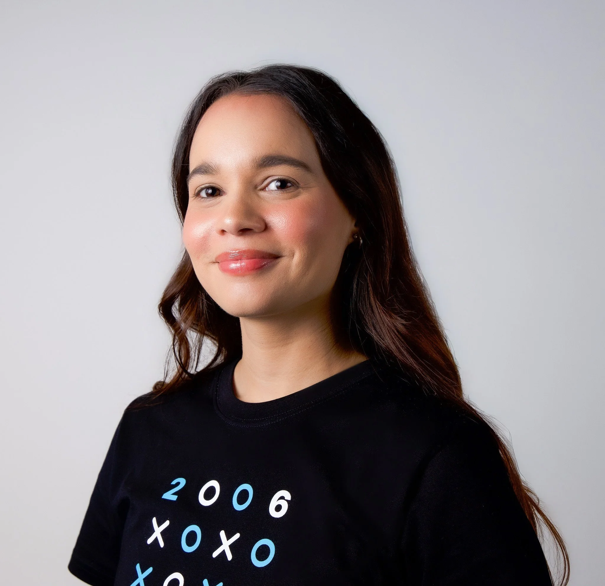 Close-up of a woman with long dark hair, smiling, wearing a black shirt with the word '2026' written on it in colorful letters, against a plain light gray background.