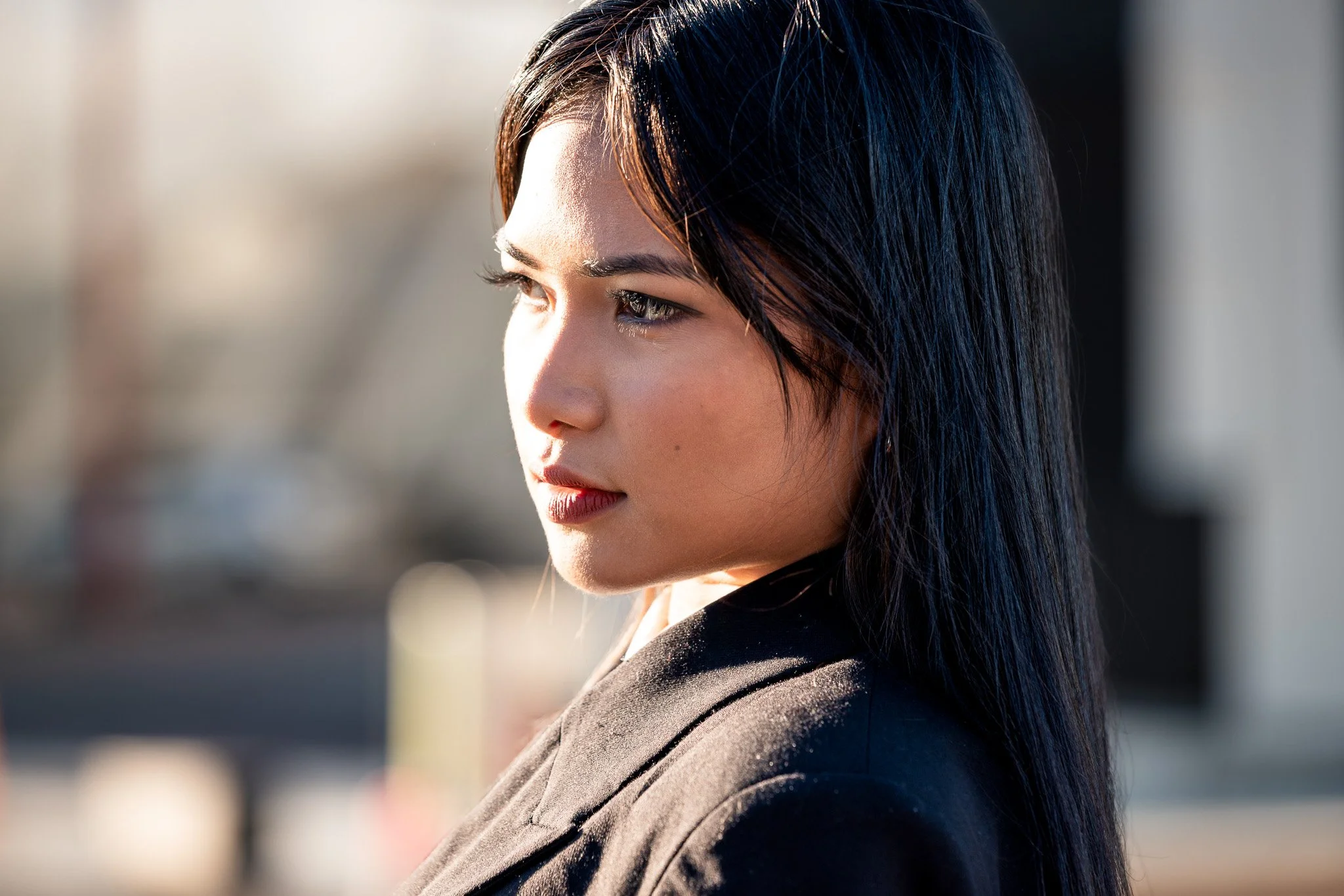 Close-up of a woman with long dark hair, looking to her right, with sunlight on her face and hair, blurred outdoor background.