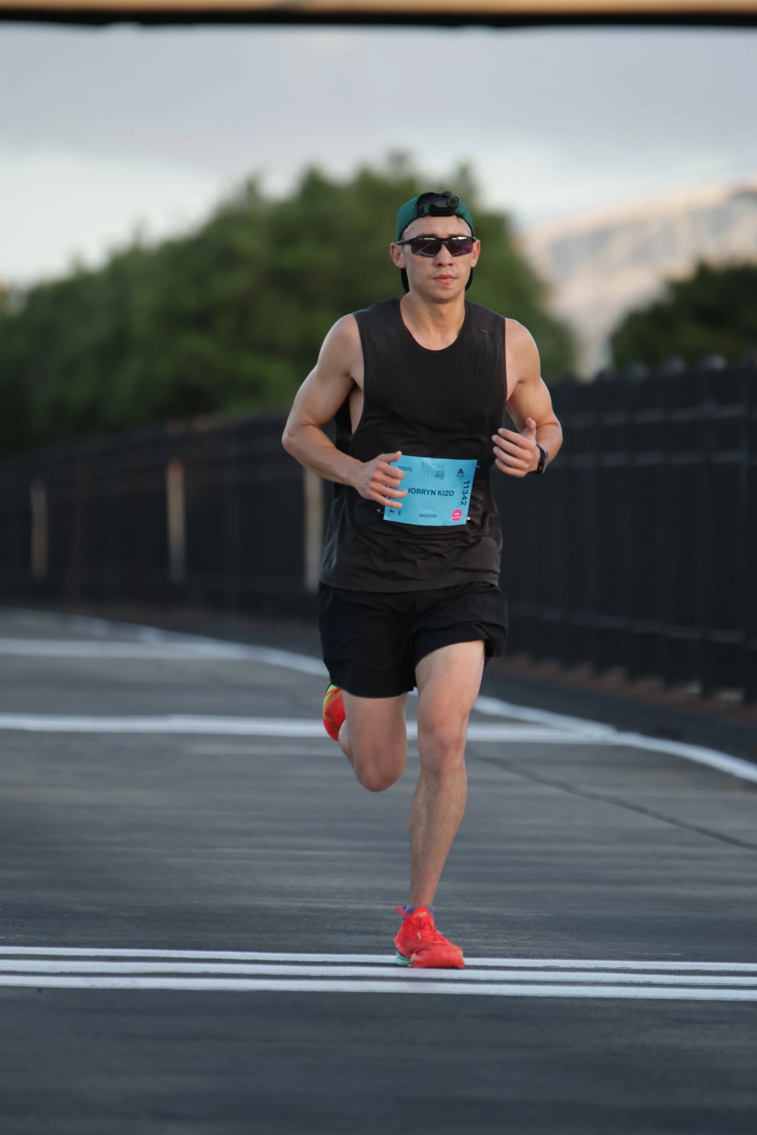 A man running in a marathon or race, wearing sunglasses, a cap with a headlamp, a black sleeveless shirt, black shorts, red running shoes, and a race bib with the name JORRYN KIZO.