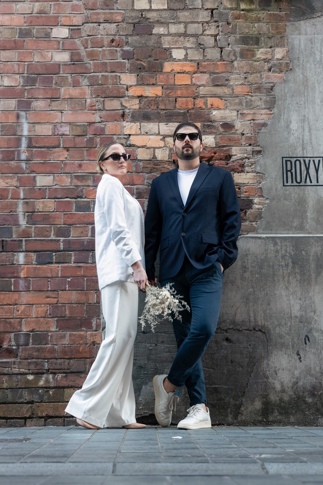 A man and woman stand against a brick wall, both wearing sunglasses. The woman is dressed in a white suit, and the man in a dark blazer and pants, with casual white sneakers. The woman holds a bouquet of dried flowers. The scene is urban and stylish.