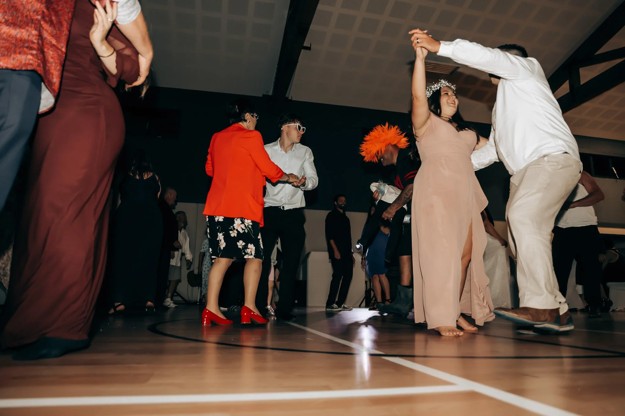 People dancing at a party or wedding reception in a gymnasium or indoor hall, with various guests dressed in formal and festive attire.