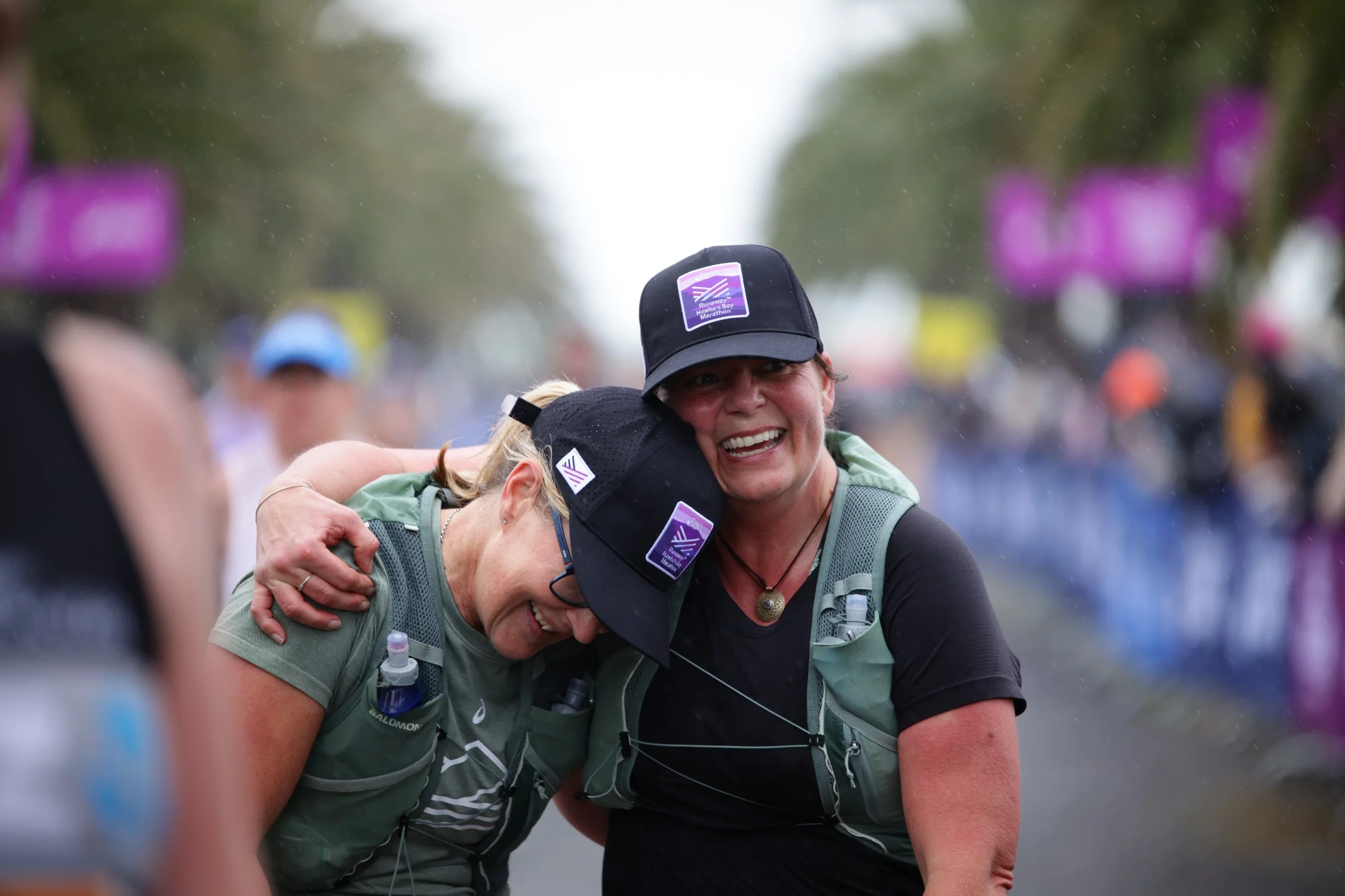 Two women hugging each other during a marathon, smiling and appearing emotional, with other participants in the background, some blurred, under a cloudy sky.