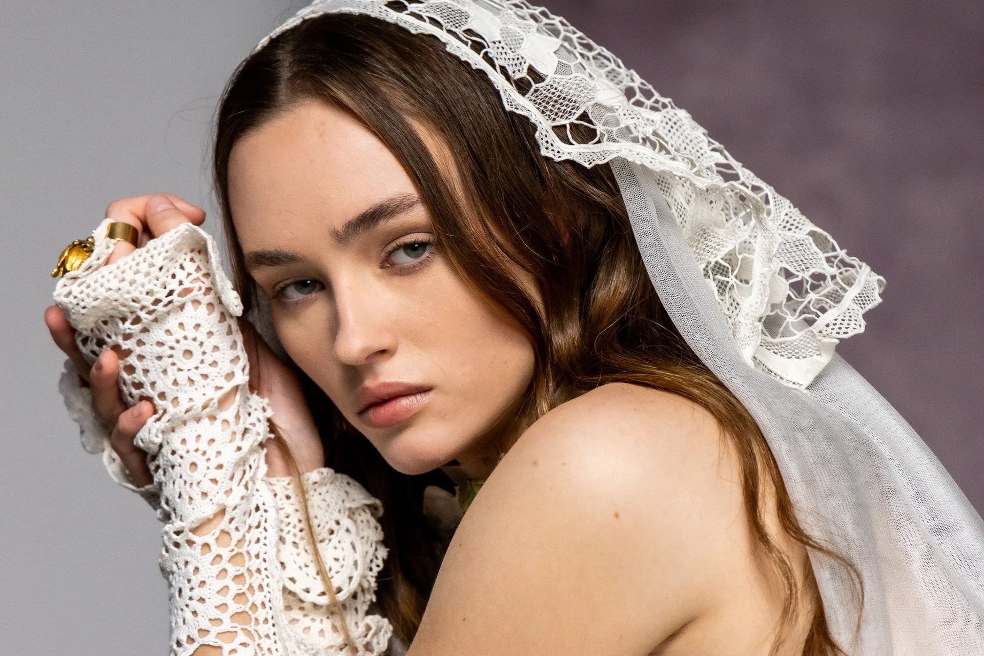 A young woman with light skin and long brown hair, wearing a white lace headdress and crochet gloves, gazes at the camera with a soft expression, against a plain gray background.