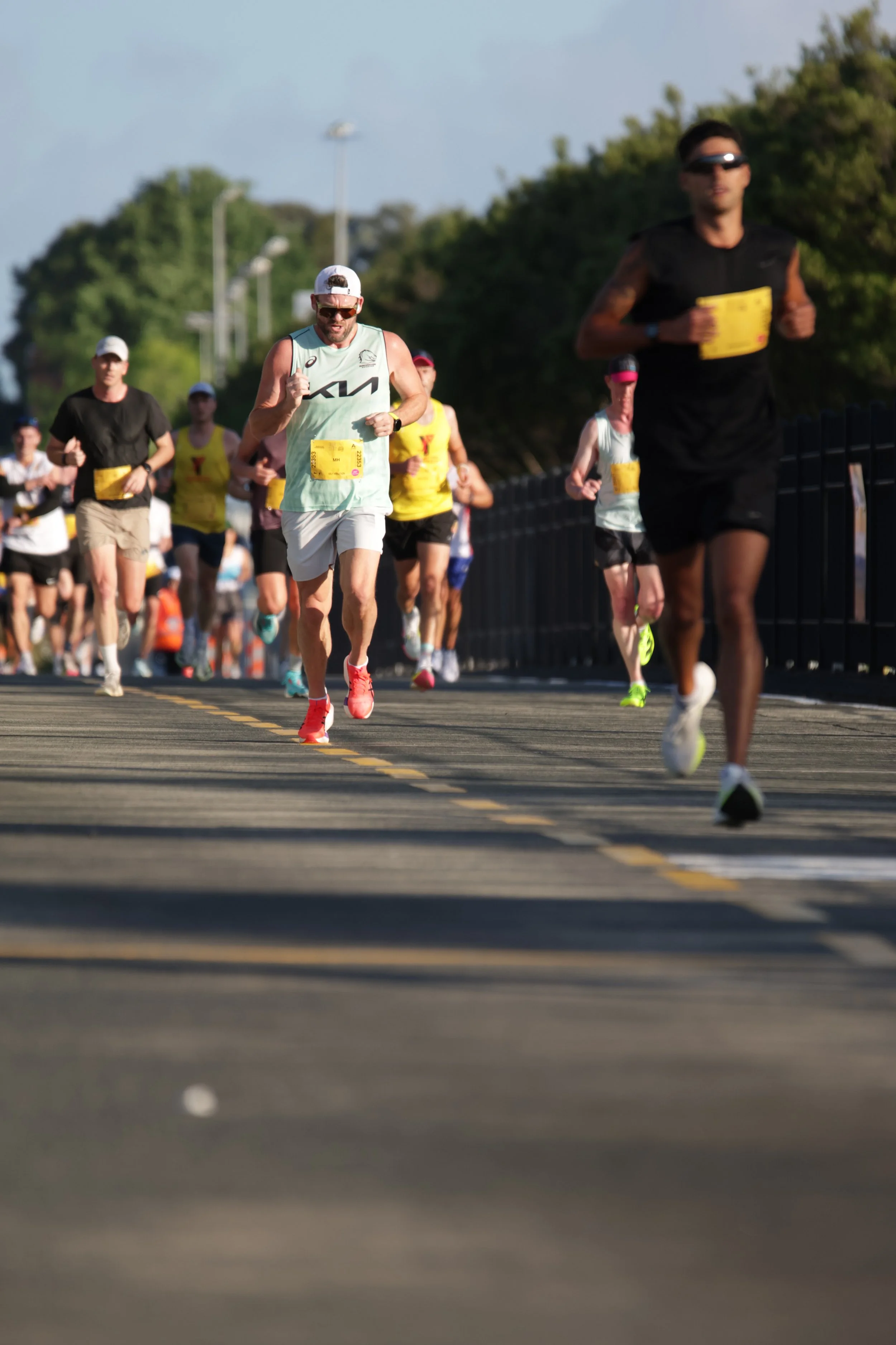 Multiple people running in a marathon outdoors on a sunny day, with trees and streetlights in the background.