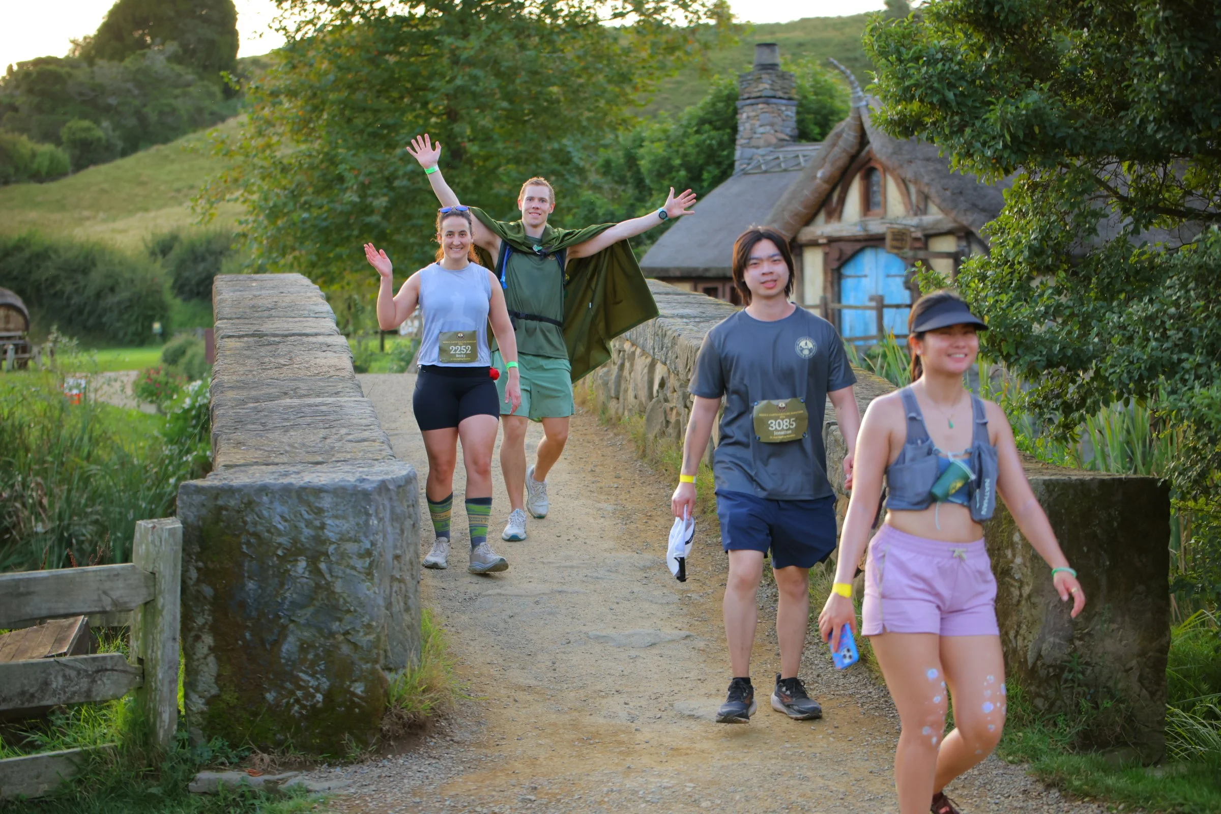 Group of people hiking on a stone bridge in a lush green landscape with a house with a thatched roof in the background.