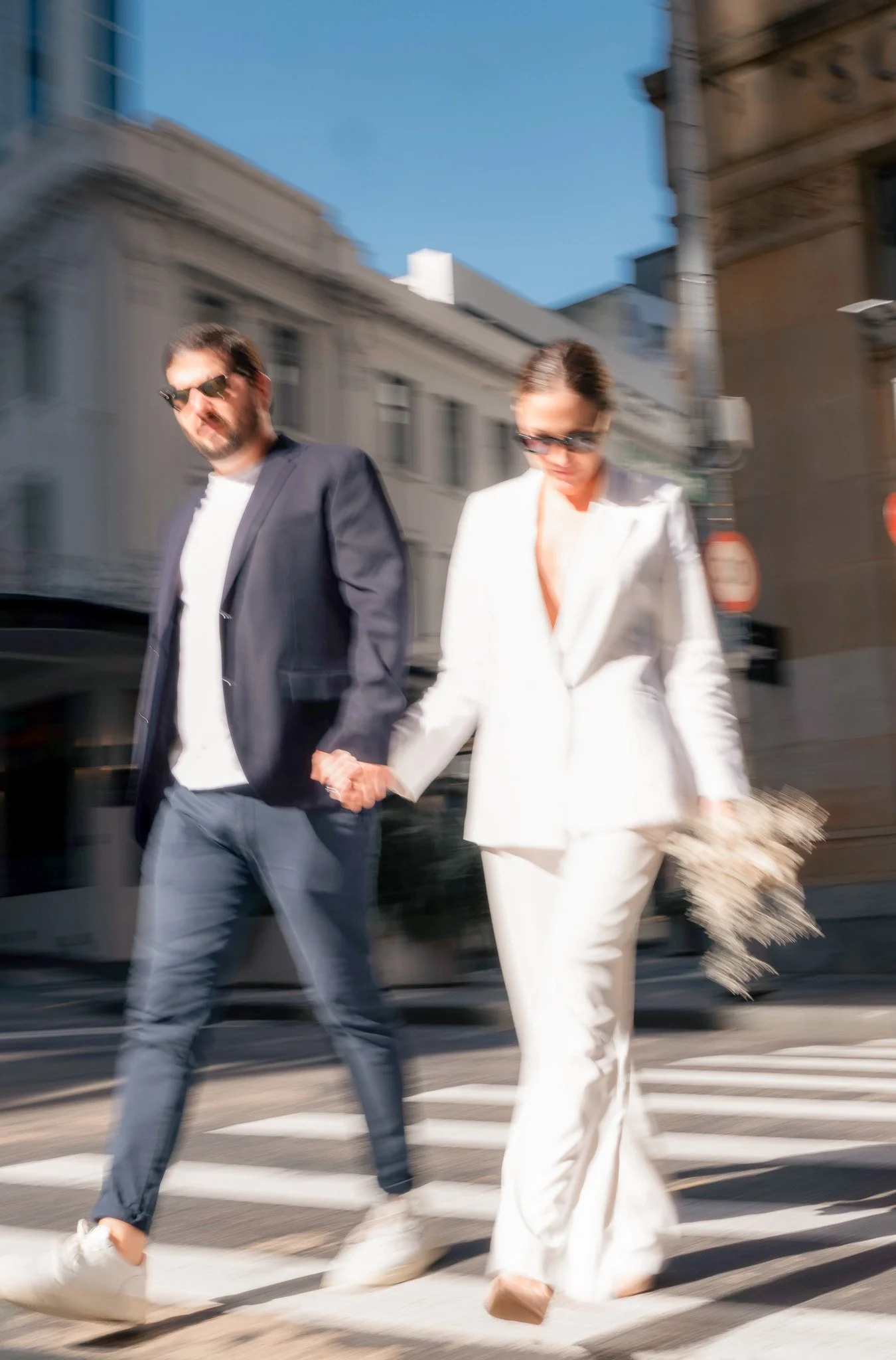 Blurred photo of a man and a woman walking hand in hand across a city street at a crosswalk. The man is wearing a dark jacket, white shirt, and sunglasses. The woman is in a white suit with wide-legged trousers, sunglasses, and is holding a fringed h