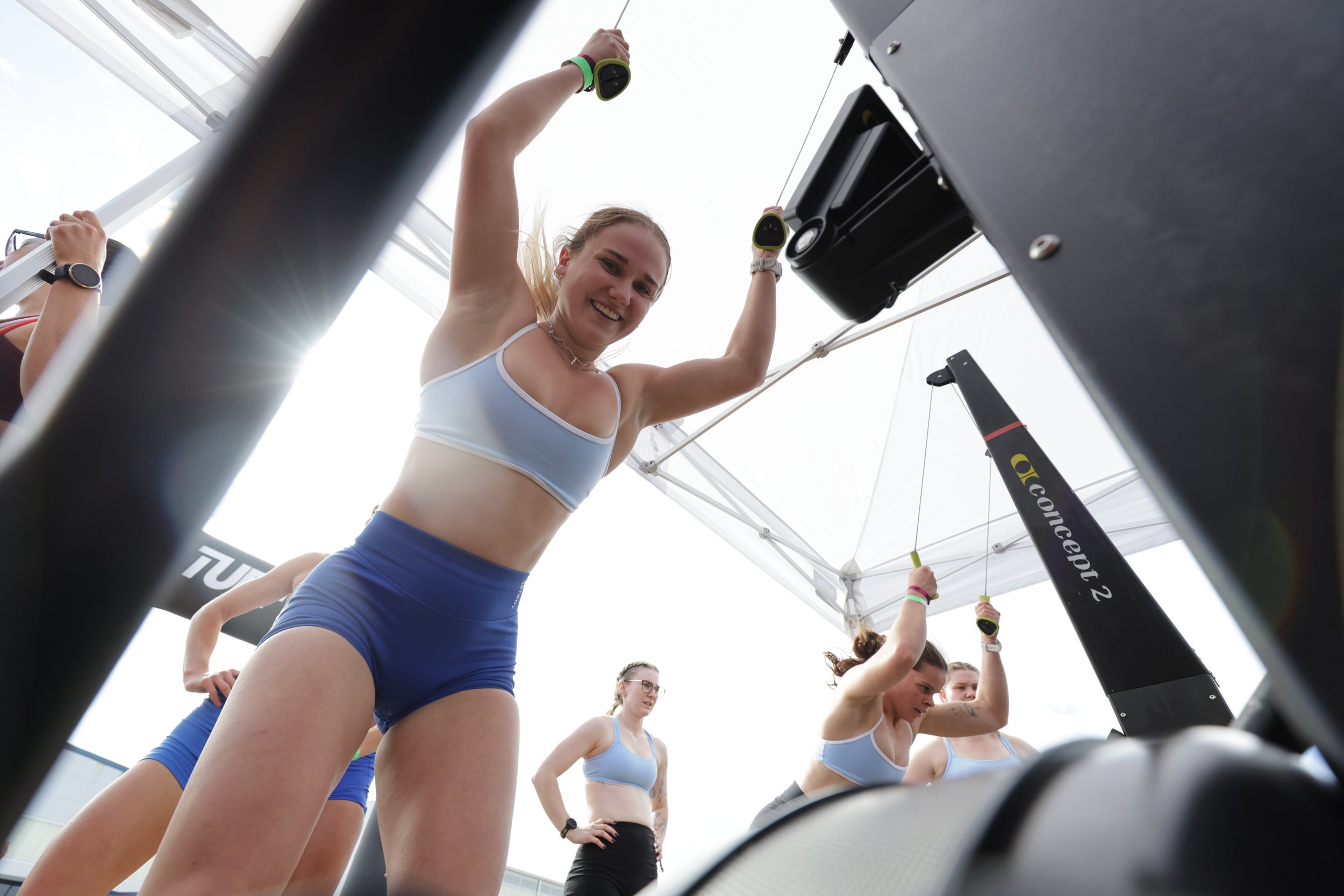 A group of young women in athletic wear standing under a white tent, with one woman smiling and holding onto a black fitness equipment frame from underneath, seen from a low angle.