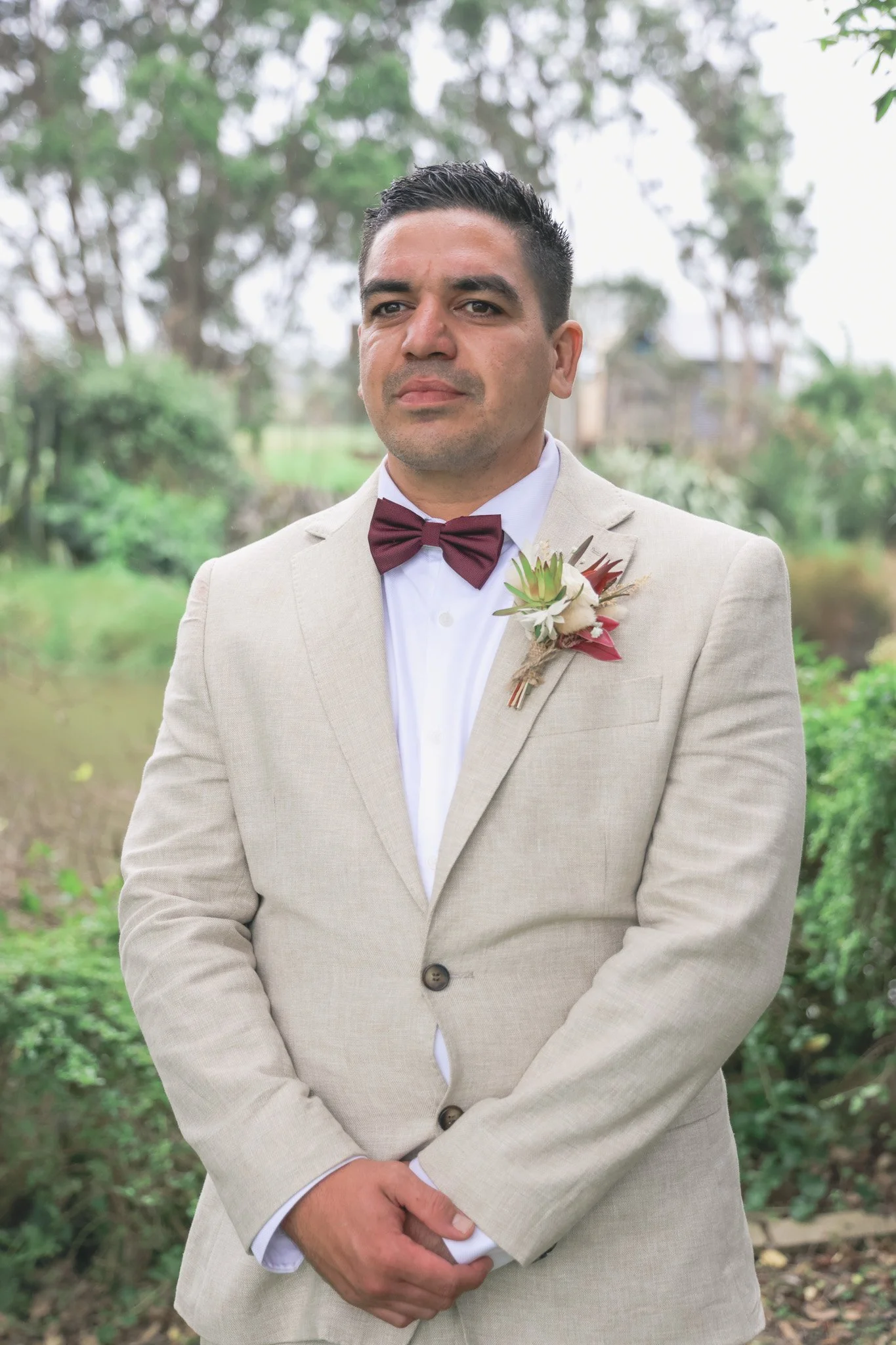 A man in a light beige suit with a burgundy bow tie and boutonniere standing outdoors, with trees and greenery in the background.