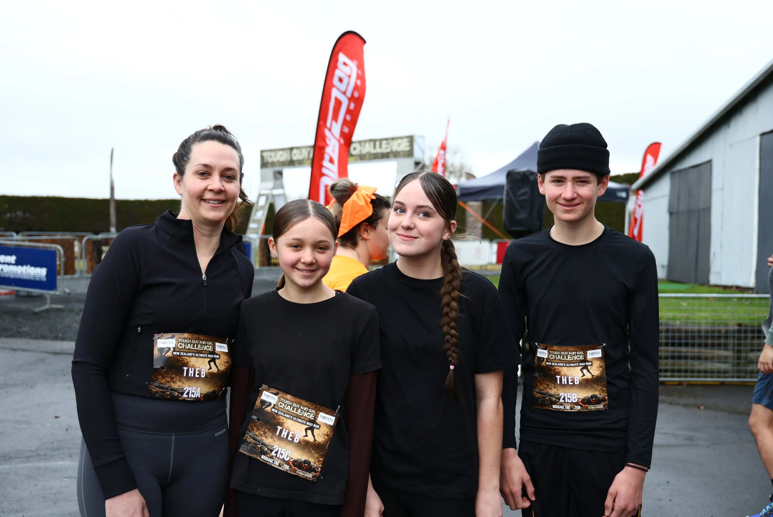 A group of four people, three girls and one woman, smiling and standing outdoors at a mud run event. They are wearing black athletic clothing with race bibs and are surrounded by race banners and tents.