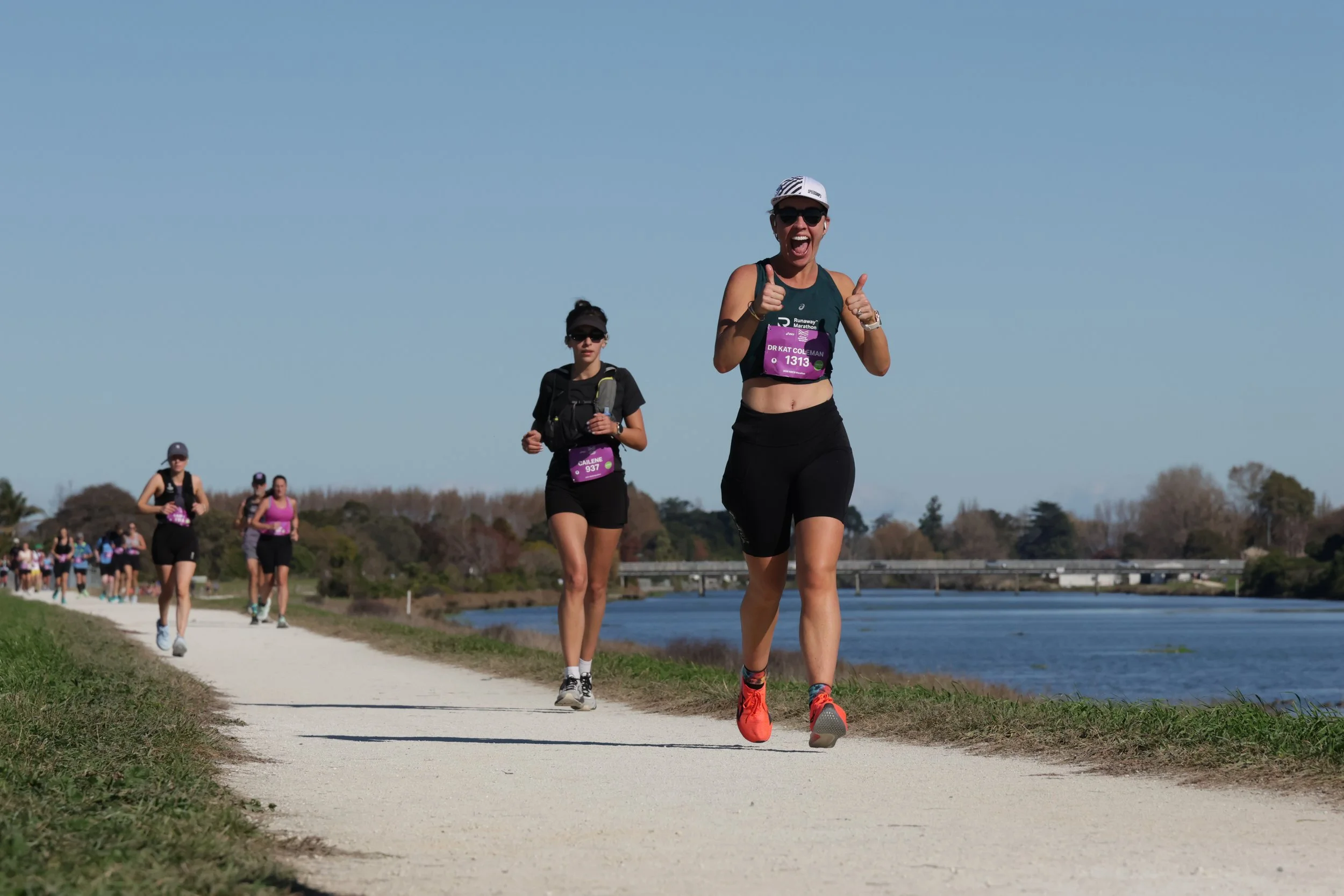 Women participating in a marathon run along a river path, wearing athletic clothing and sunglasses, with a bright blue sky overhead and trees in the background.