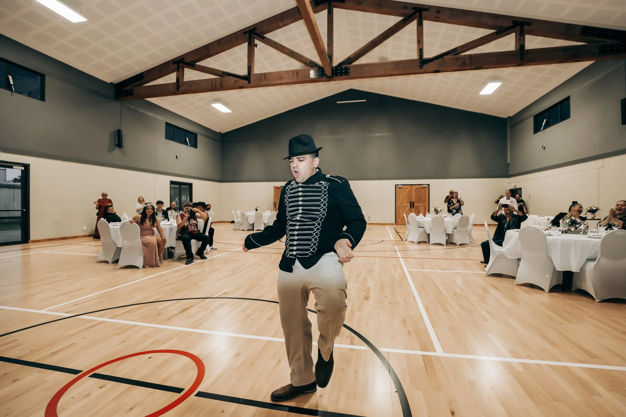 A man dressed in a military-style jacket, beige pants, and a black hat dances in the center of a large indoor event space with wooden beams on the ceiling and several round tables with seated guests around him.