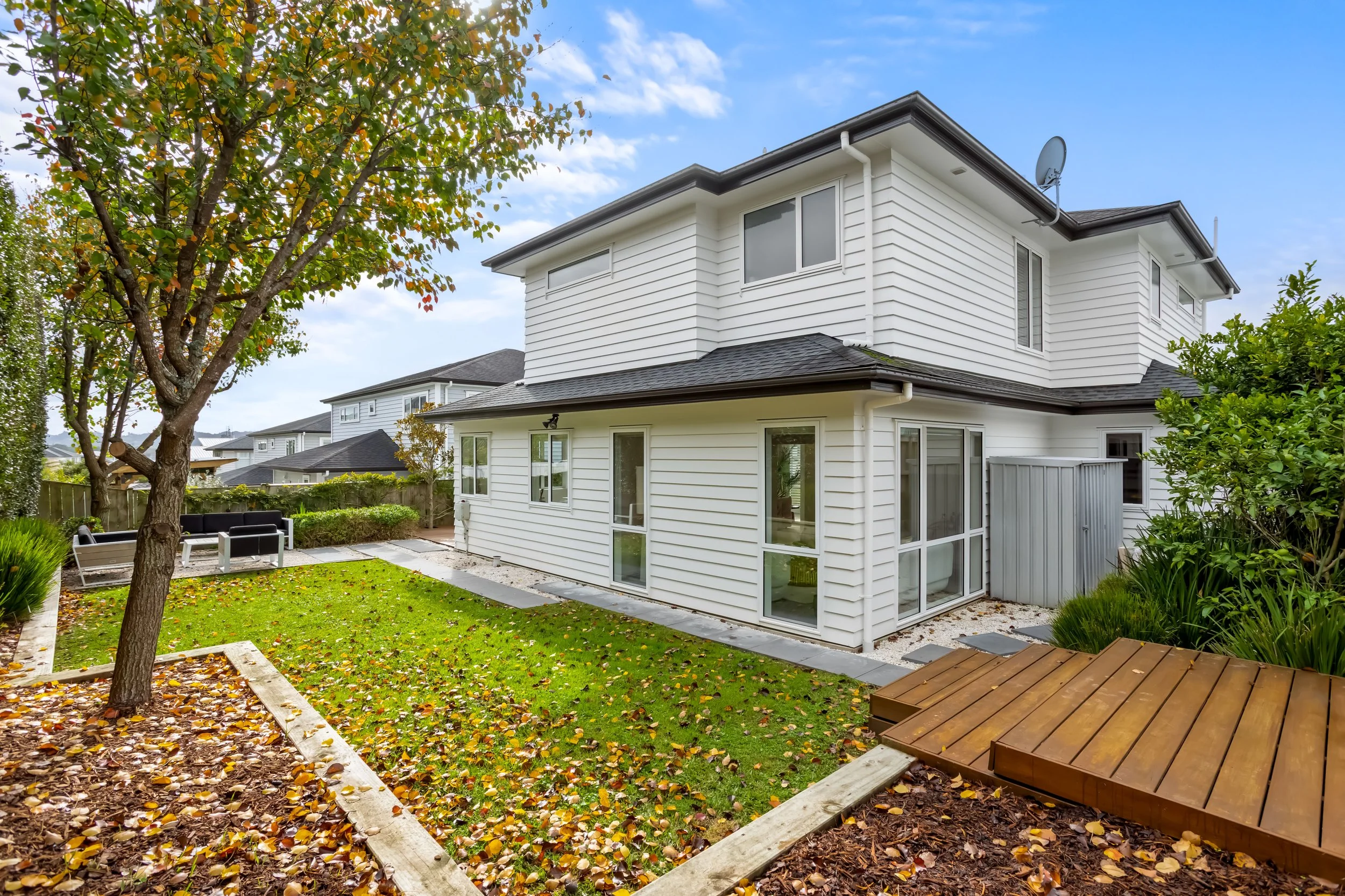 Backyard with green lawn, tree with autumn leaves, wooden deck, and a white two-story house with multiple windows and a satellite dish.