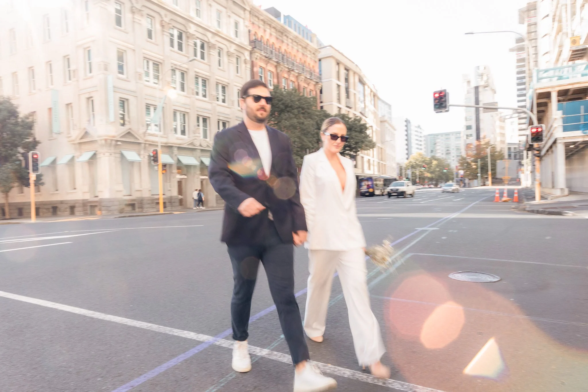 A man and woman walking across a city street during daytime, with tall buildings in the background and traffic lights visible.