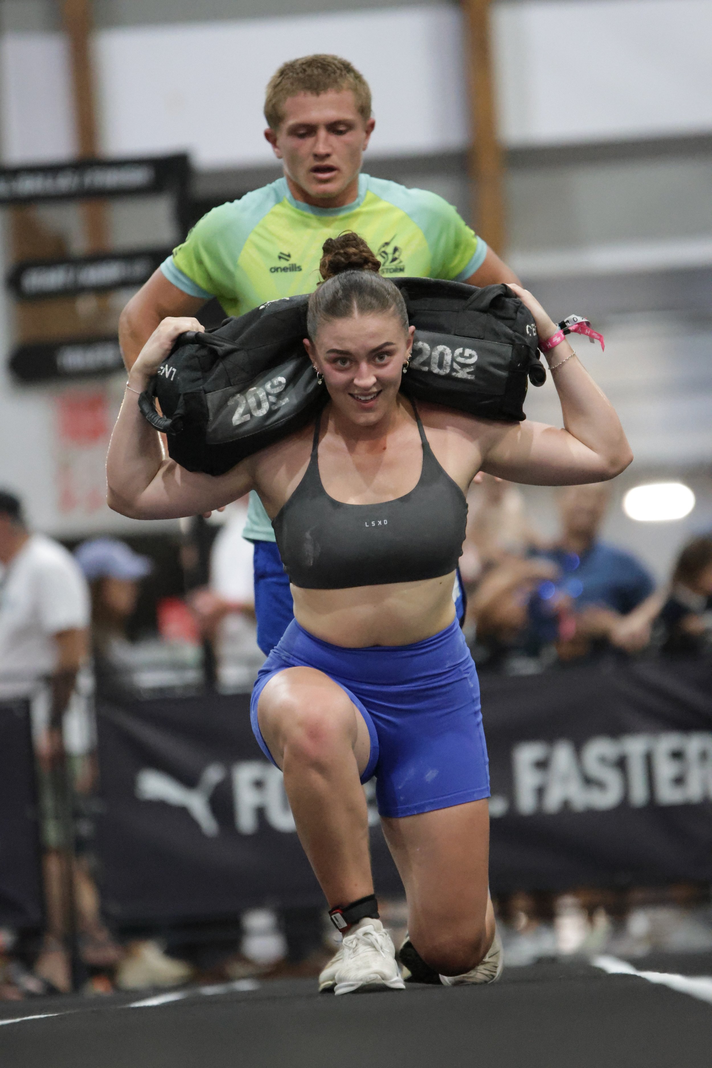 A young woman with a braid is running indoors while carrying a large black duffel bag on her shoulders. A young man in a green shirt is running behind her. The background shows a busy athletic facility with other people and workout equipment.
