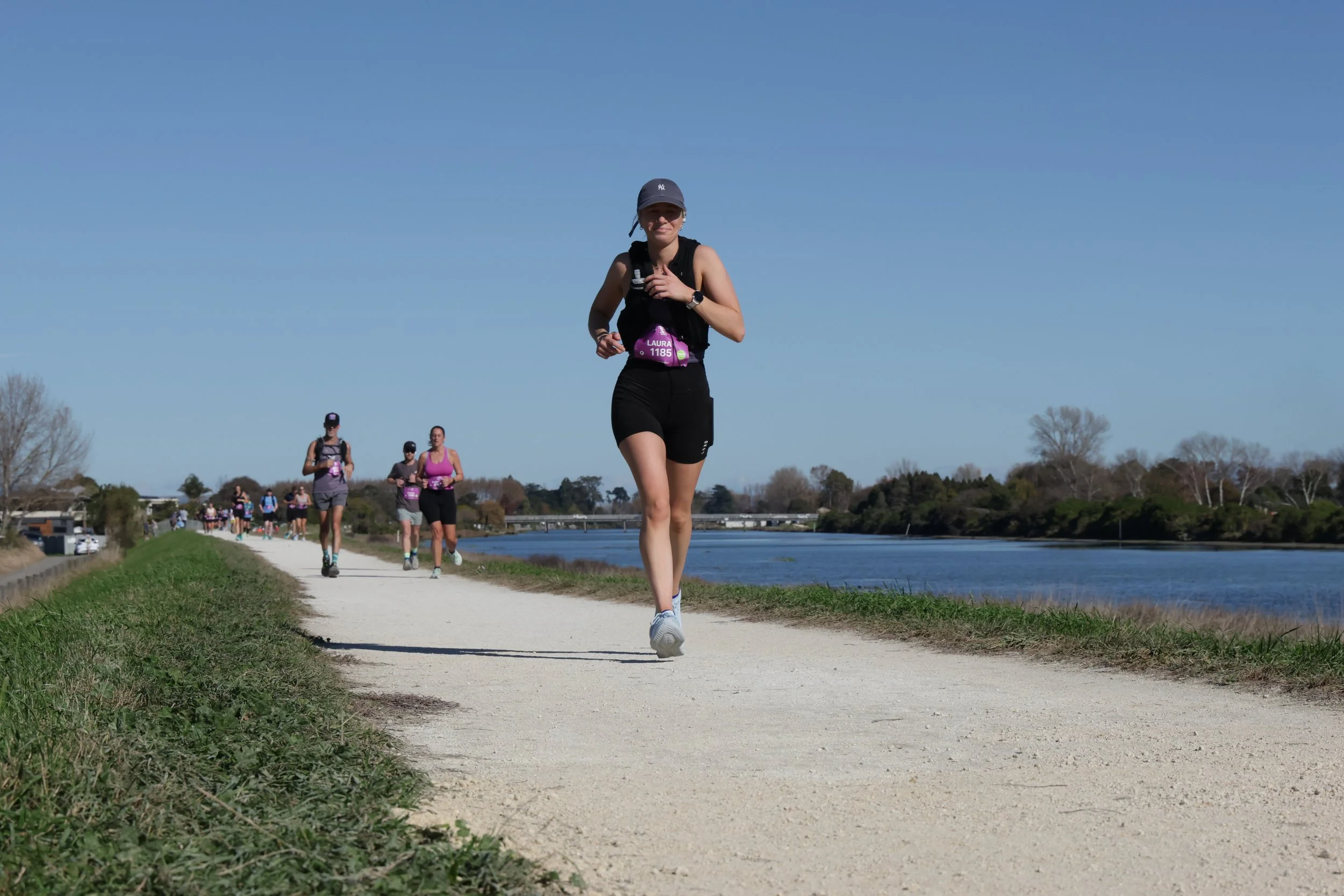 Women running on a dirt pathway next to a river on a sunny day, with others running behind her and a tree-lined landscape.