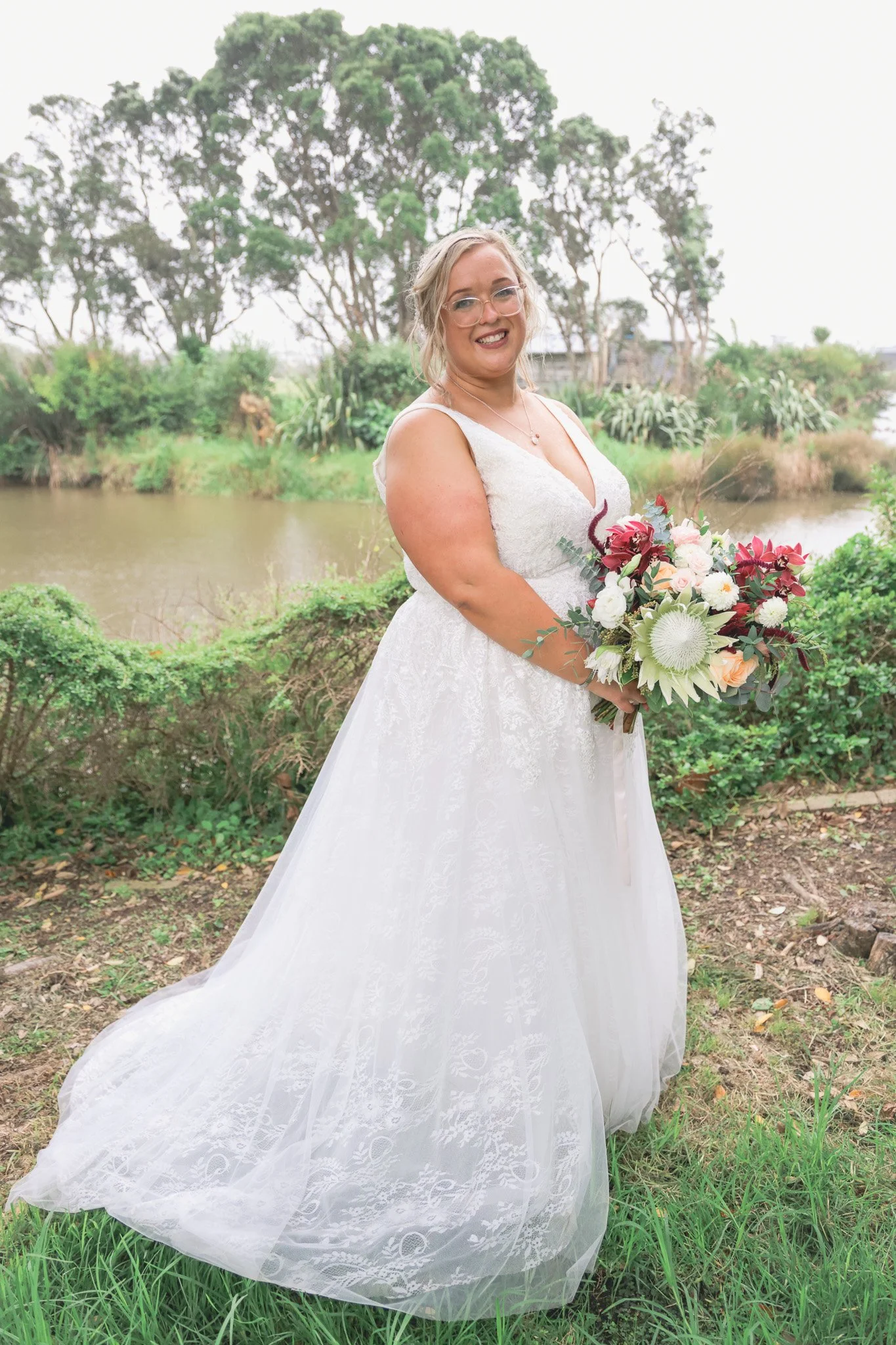 A woman in a white wedding dress holding a bouquet of flowers outdoors by a river with green trees in the background.
