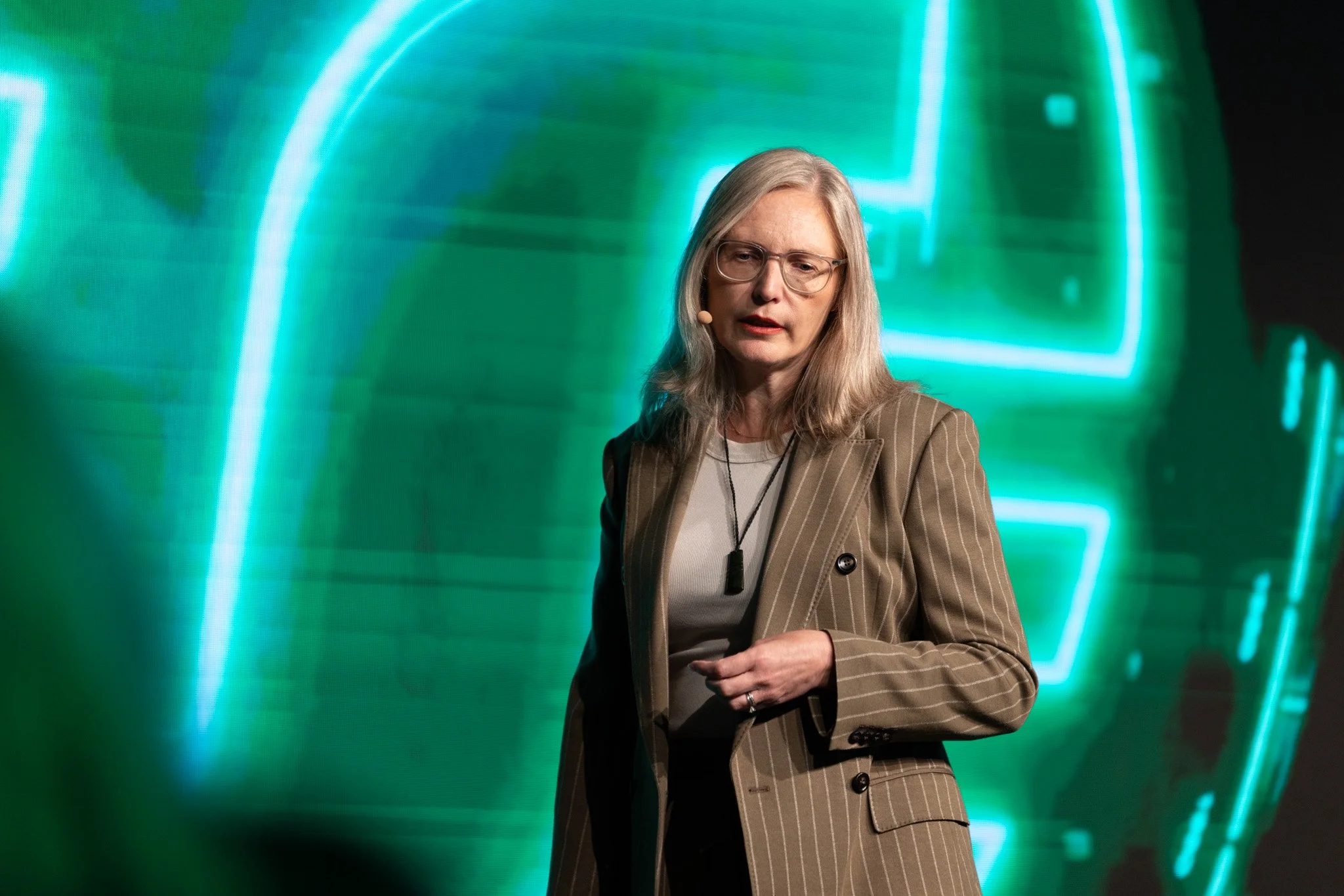 A woman with gray hair and glasses speaking at a tech conference, standing in front of a green digital backdrop.