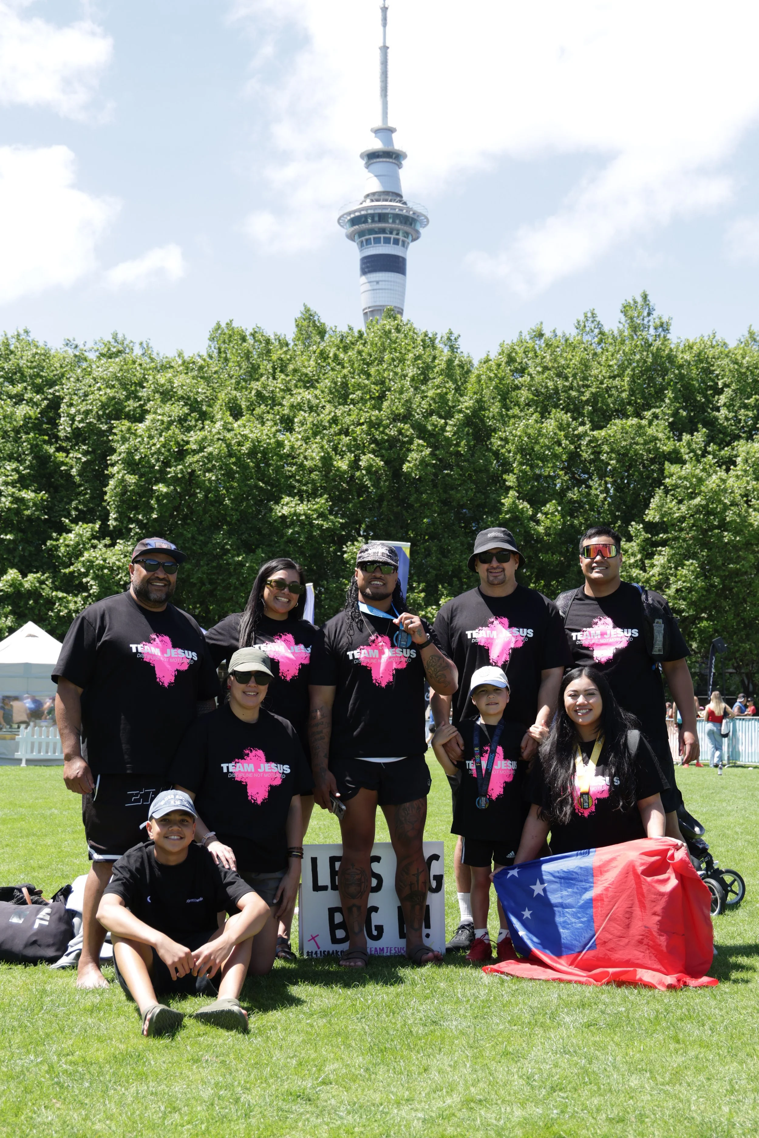 Group of diverse people wearing black t-shirts with pink writing, standing on grass in front of green trees, with a television tower in the background and the sky above. Some holding signs and flags. It appears to be an outdoor rally or demonstration