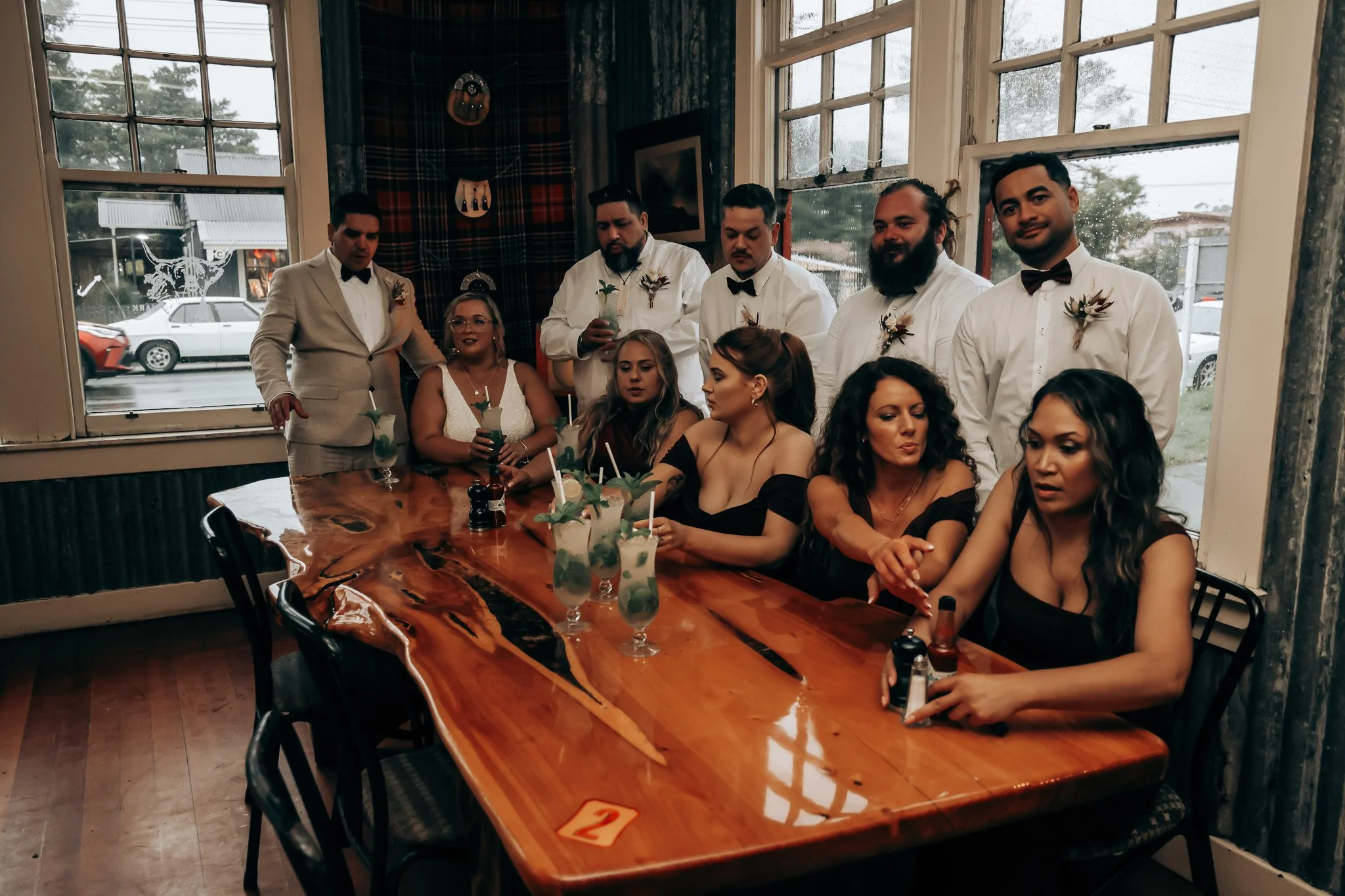 A group of nine people, dressed in formal attire, sitting around a wooden table in a restaurant or banquet hall. Five of them are standing behind the table, and four are seated. The table has multiple tropical drinks with umbrellas and straws. Large 