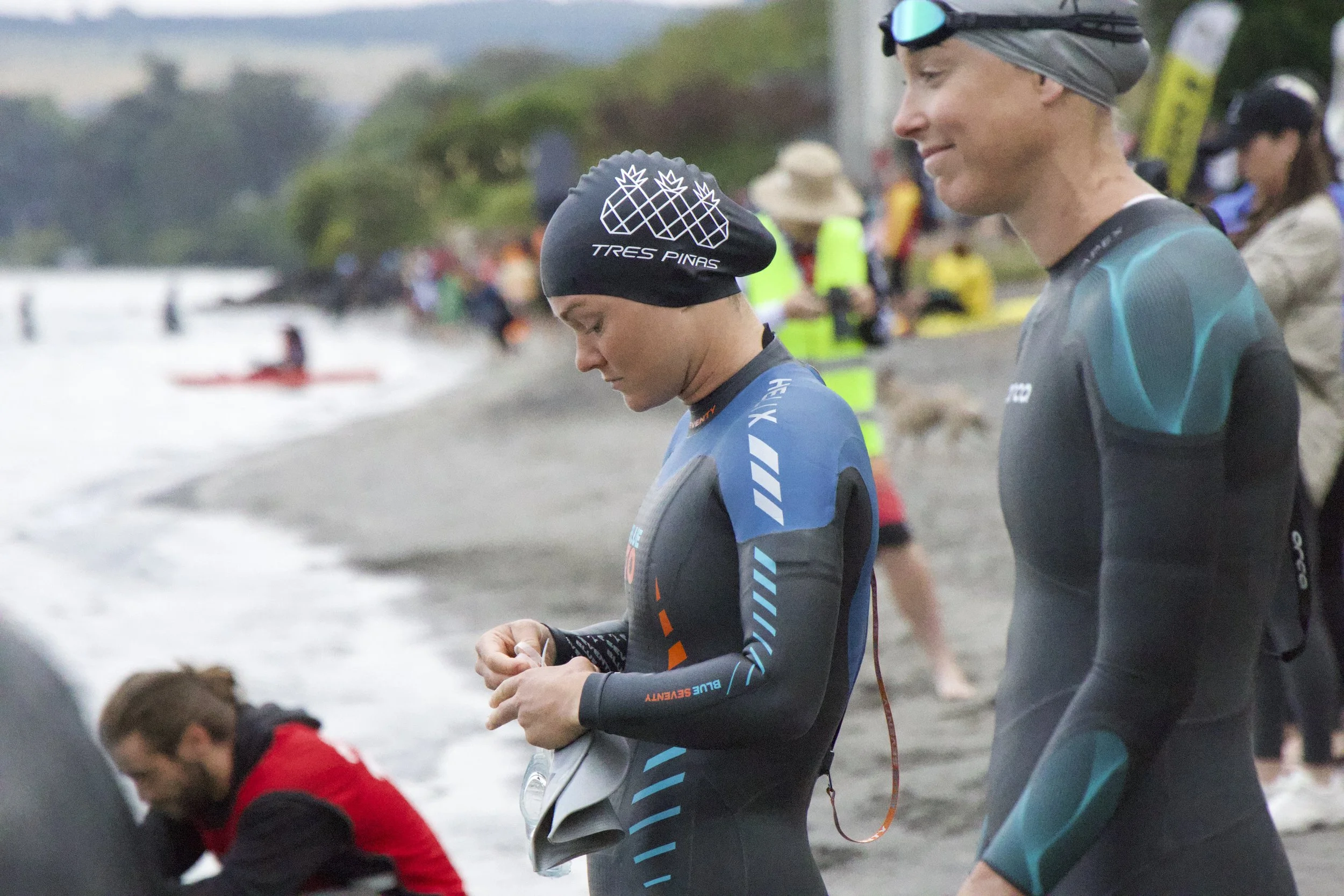 Two athletes dressed in wetsuits are standing on a beach near the water, with other people and kayaks visible in the background, likely preparing for a triathlon or swim event.