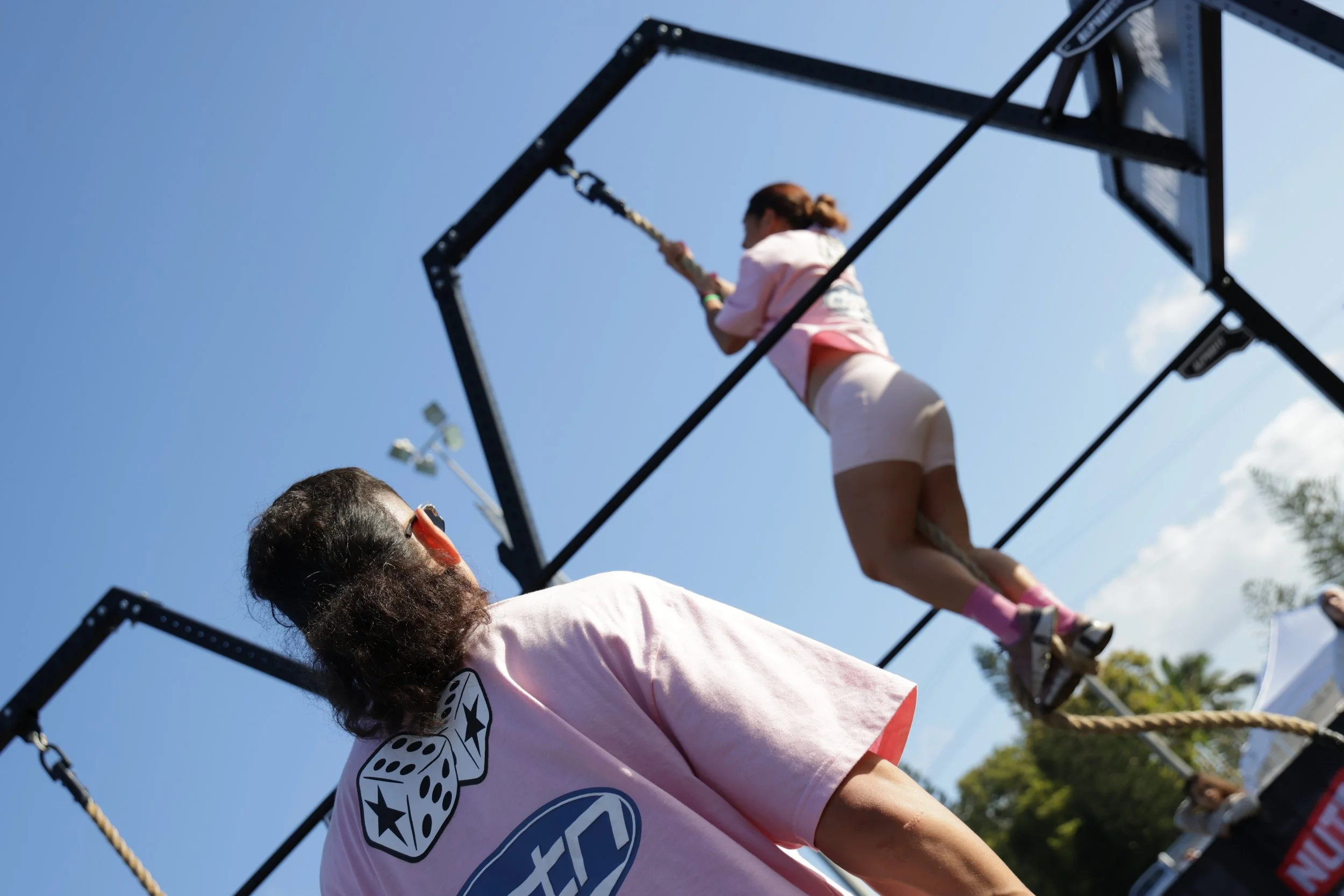 A person with reddish hair and a pink shirt is balancing on someone's shoulders during an outdoor event, with a blue sky and trees in the background.