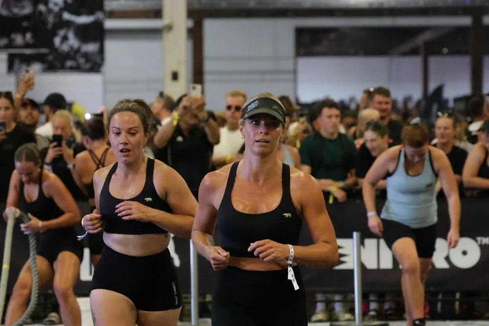 Women participating in a fitness event indoors, dressed in athletic wear, with spectators watching in the background.