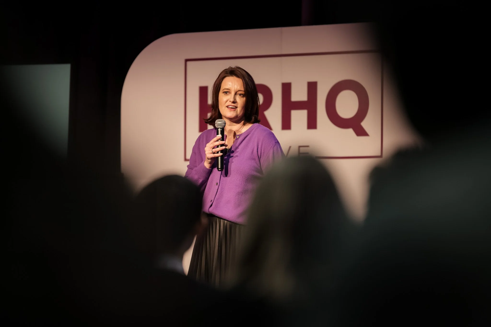 Woman speaking into a microphone on stage at an event with a large RHQ logo in the background.