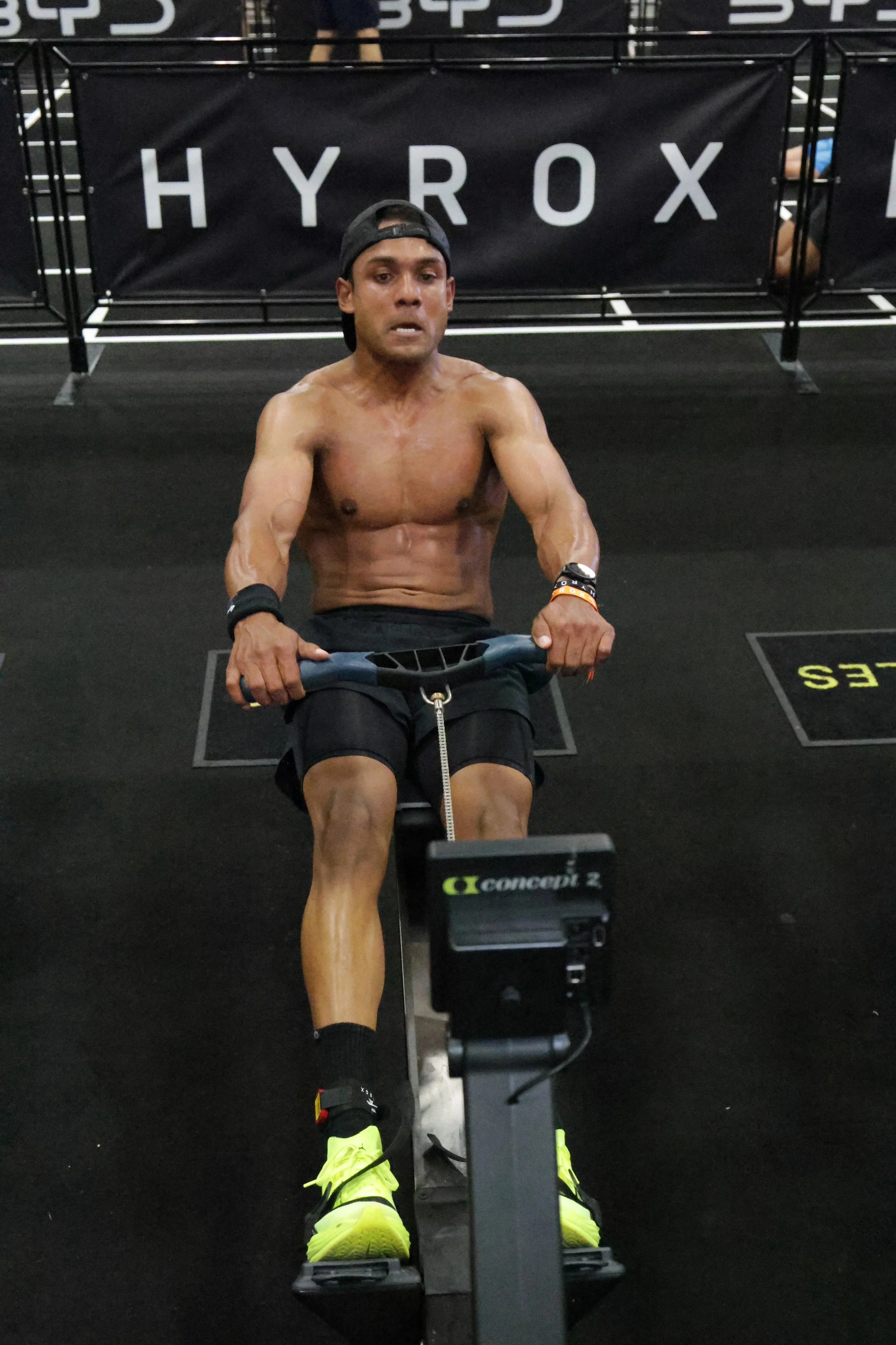 A man exercising on a rowing machine at a gym with black flooring and a black banner with white text reading 'HYROX' in the background.