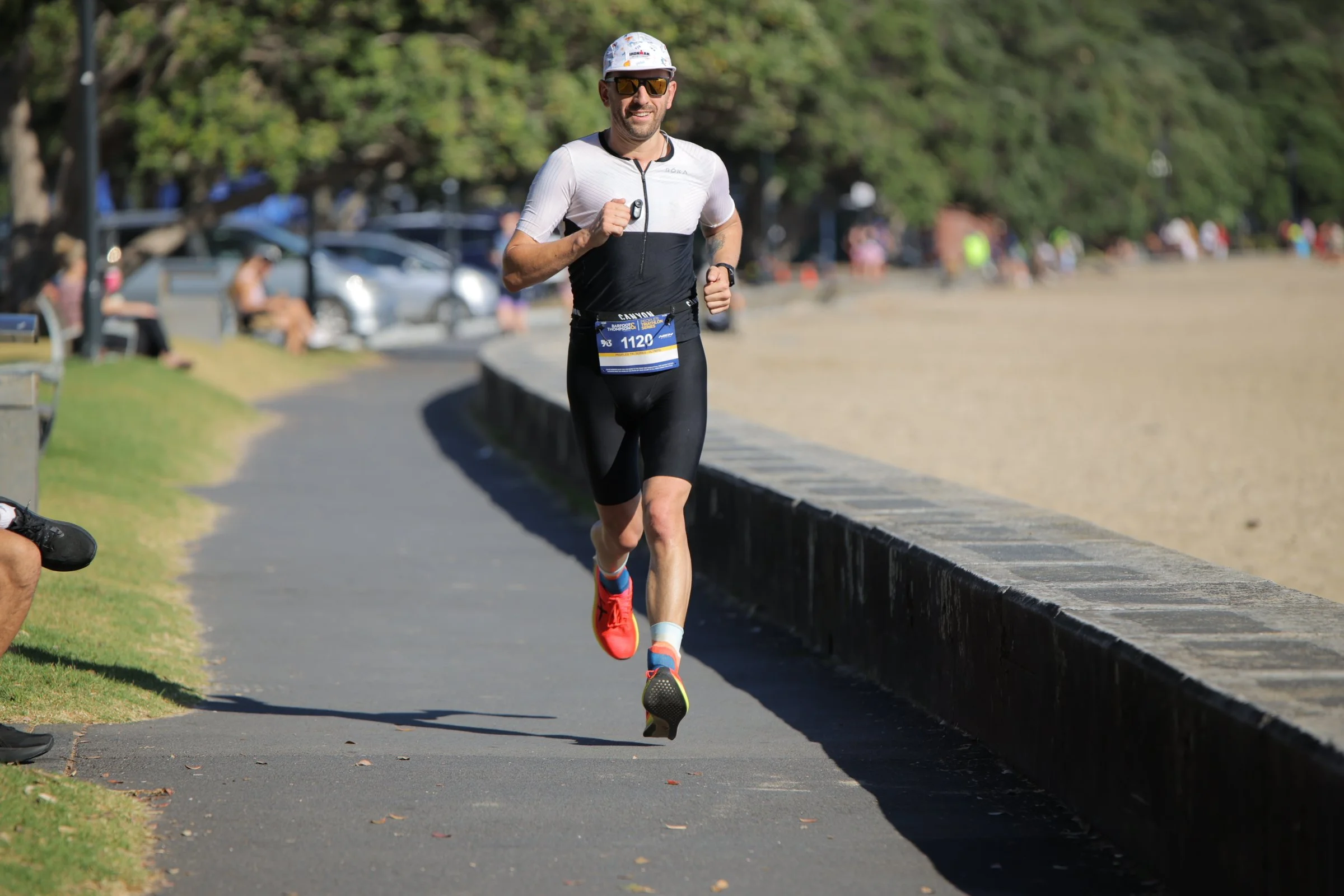 A man running in a park during a race, wearing a white and black athletic outfit, red running shoes, sunglasses, and a race bib number 1120. He is smiling and appears to be enjoying the run. In the background, there are trees, parked cars, and specta