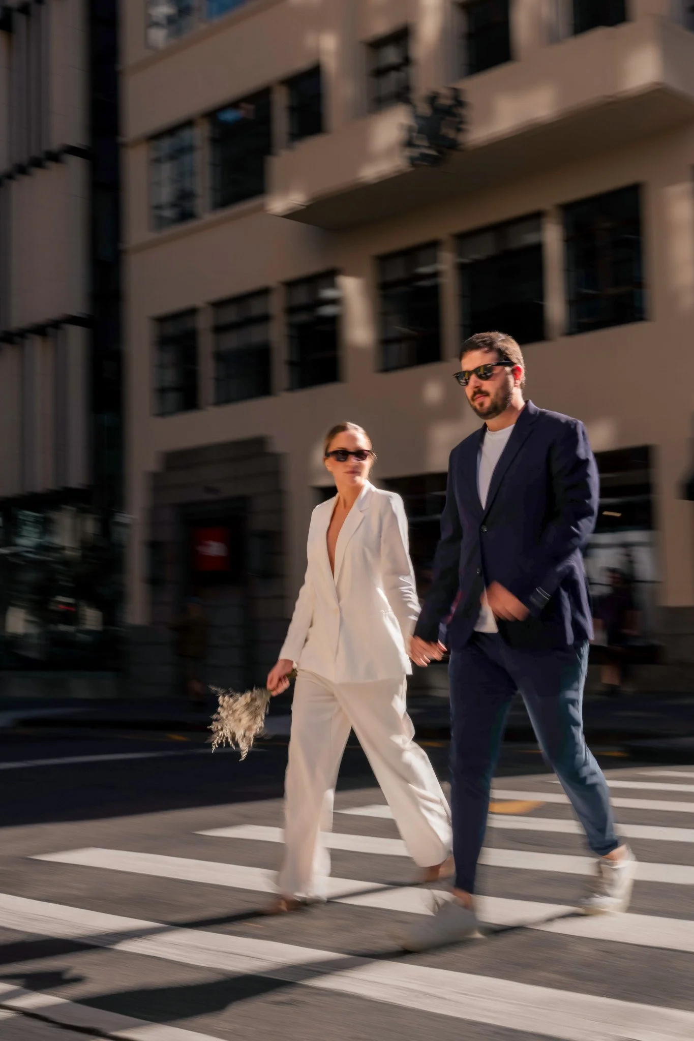 A man and woman walking hand in hand across a city street crosswalk, both dressed stylishly in suits and wearing sunglasses, with tall buildings in the background.