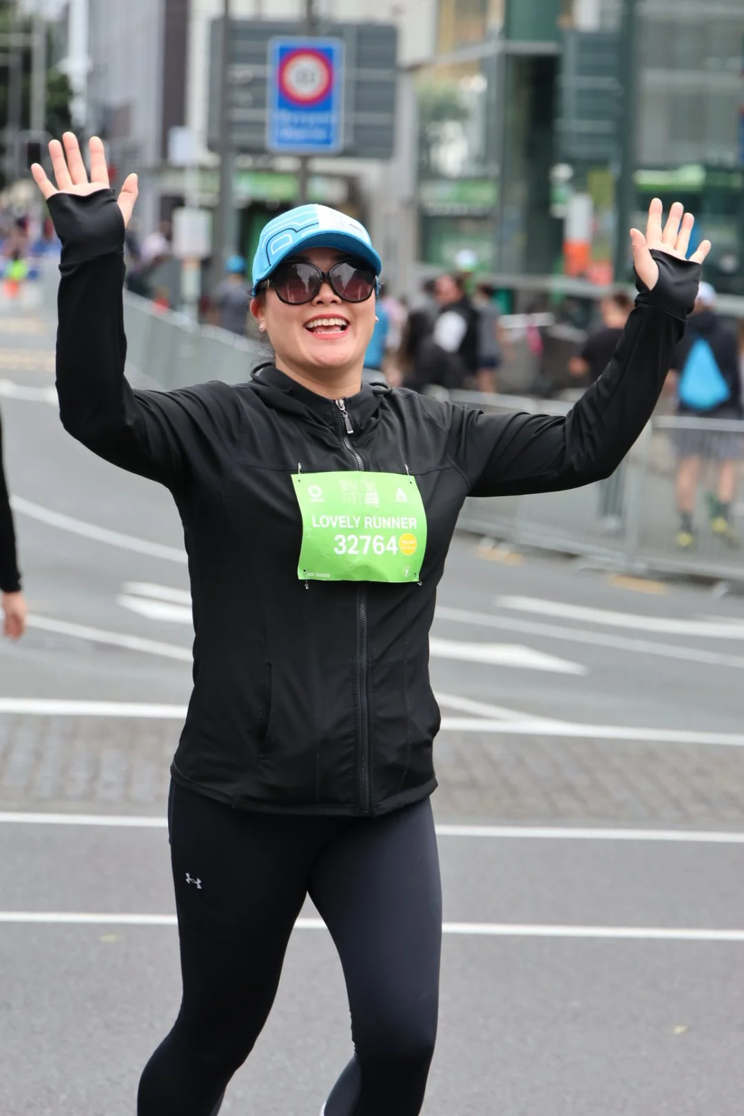 A woman in running gear, wearing sunglasses and a blue cap, smiling and waving at a marathon event. She has a green race bib labeled 'LOVELY RUNNER 32764' attached to her black jacket. The background shows other runners and city buildings.