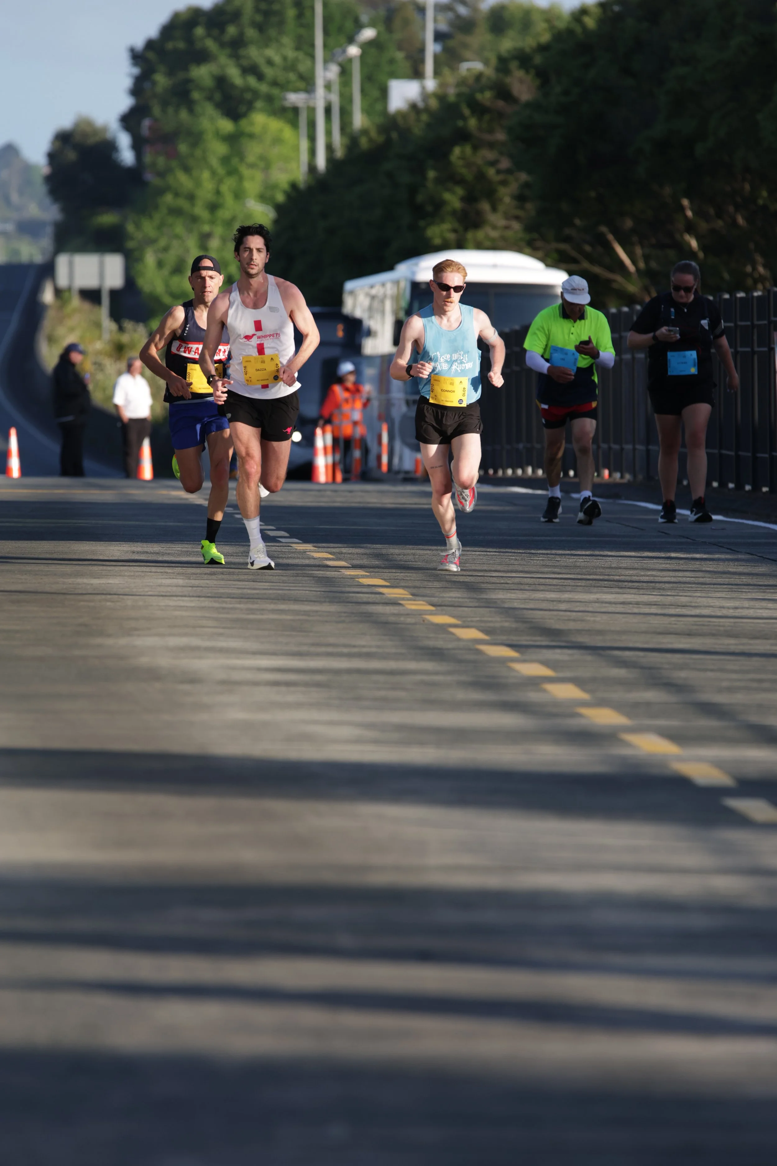 A group of five runners participating in a marathon on a paved road, with some spectators and officials in the background, surrounded by green trees and a bus.