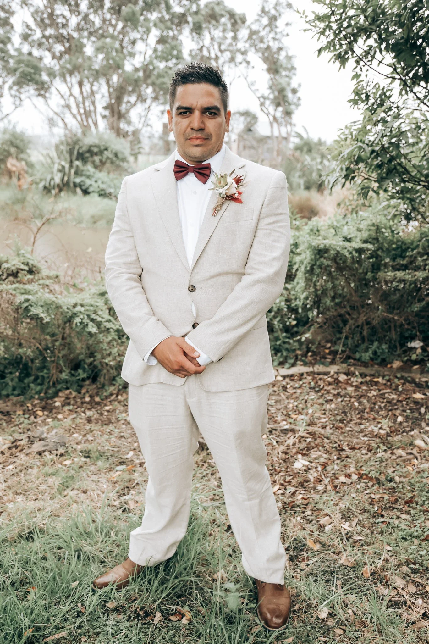 Man in a light-colored suit with a red bow tie and boutonnière standing outdoors in a natural setting with trees and grass.