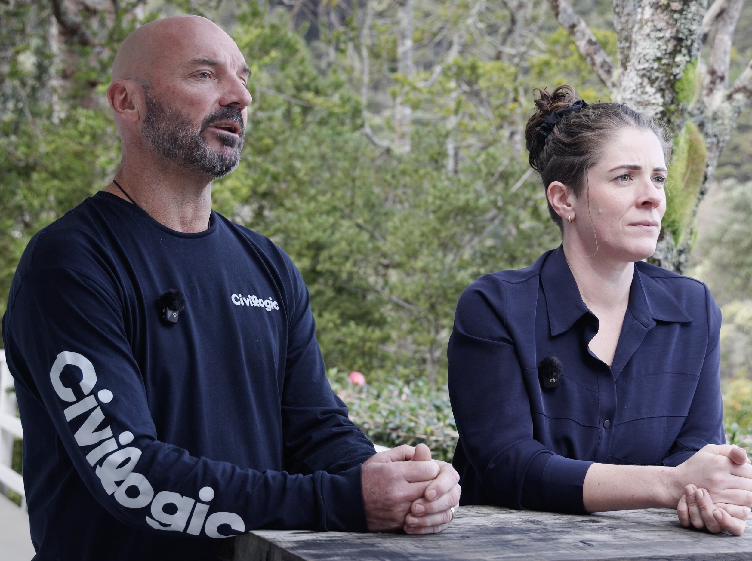 A man and a woman sitting outdoors at a table, with trees and greenery in the background. The man has a bald head and is wearing a navy blue shirt with white text, and the woman has dark hair tied back with a black hairband, wearing a dark shirt. Both are clasping their hands together on the table.