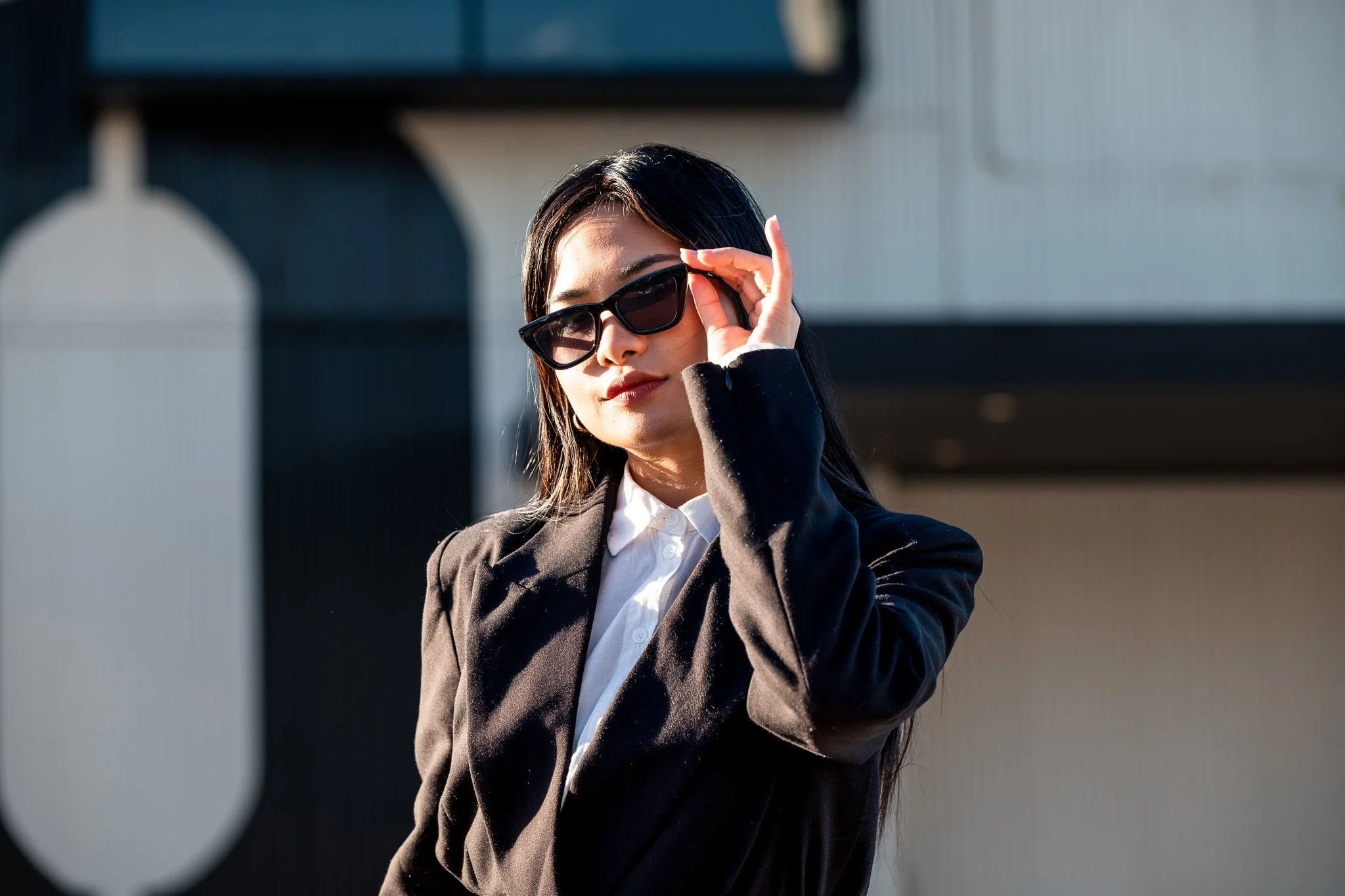 A woman with long dark hair wearing large black sunglasses, a black blazer, and a white shirt, standing outdoors against a modern building. She is adjusting her sunglasses with her right hand.
