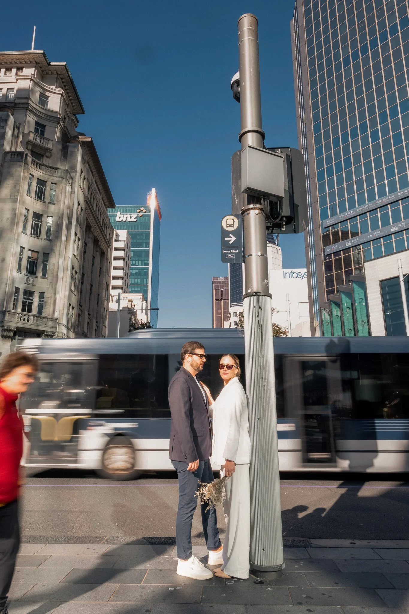 Two people, a man in a dark suit and a woman in a white outfit, standing close together and holding hands near a street corner with a bus passing by. There is a tall street pole with a sign pointing right to Lower Albert, 150 meters, and a bus stop i