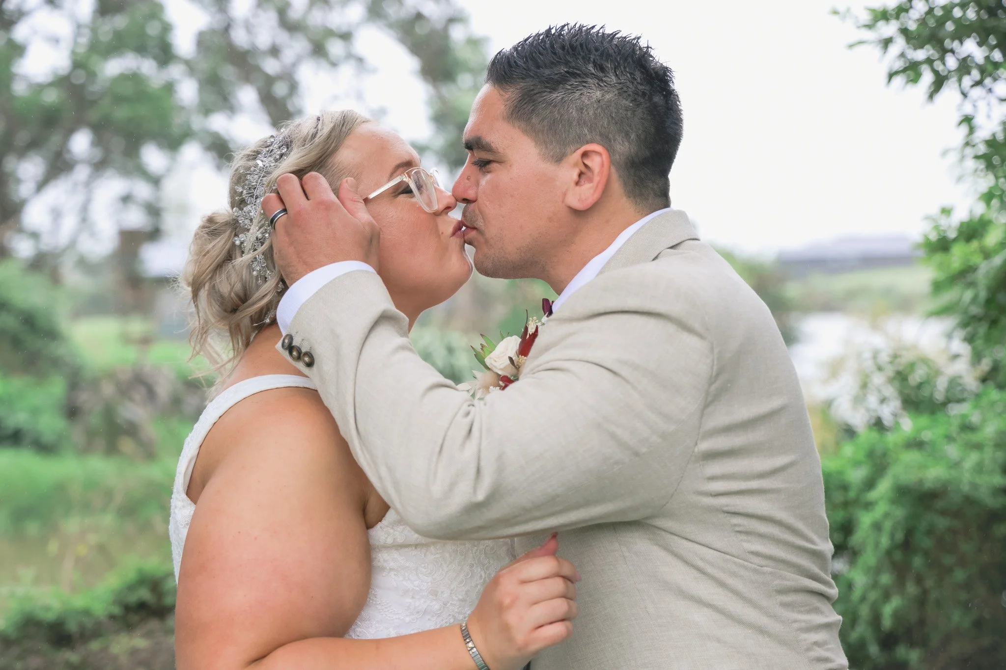 A bride and groom sharing a kiss outdoors during their wedding, with the groom gently holding the bride's face and the bride holding the groom's hand.