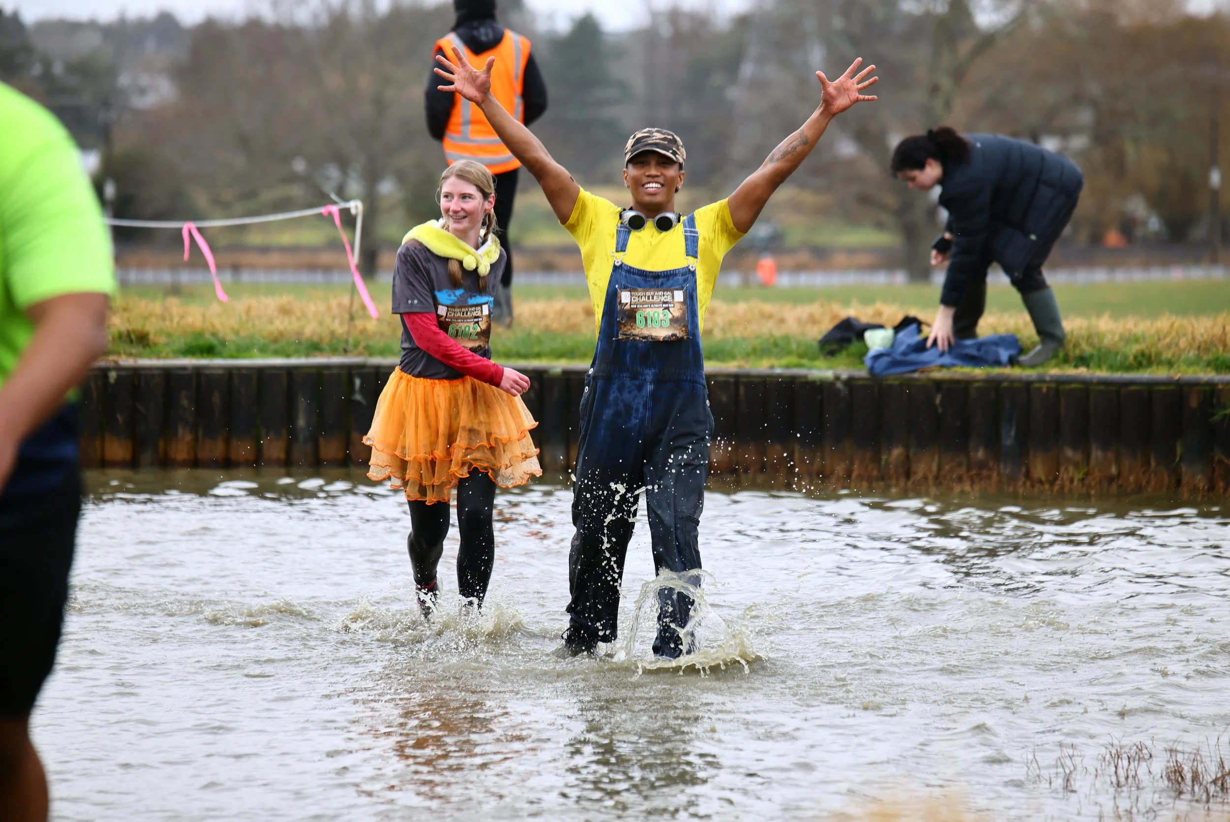 People participating in a race or outdoor event crossing a shallow body of water, with some smiling and raising their arms, while others are preparing or helping nearby.