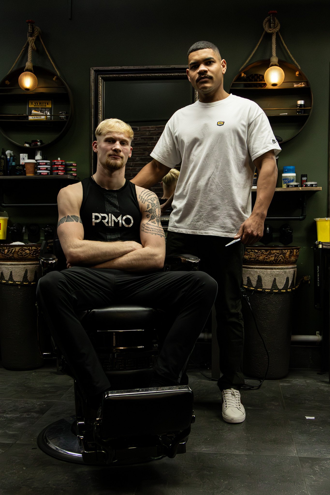 Two men in a barbershop, one sitting in a barber chair with crossed arms and the other standing beside him. The seated man has tattoos and a black tank top, while the standing man has a short haircut, beard, white T-shirt, and white sneakers. The bac