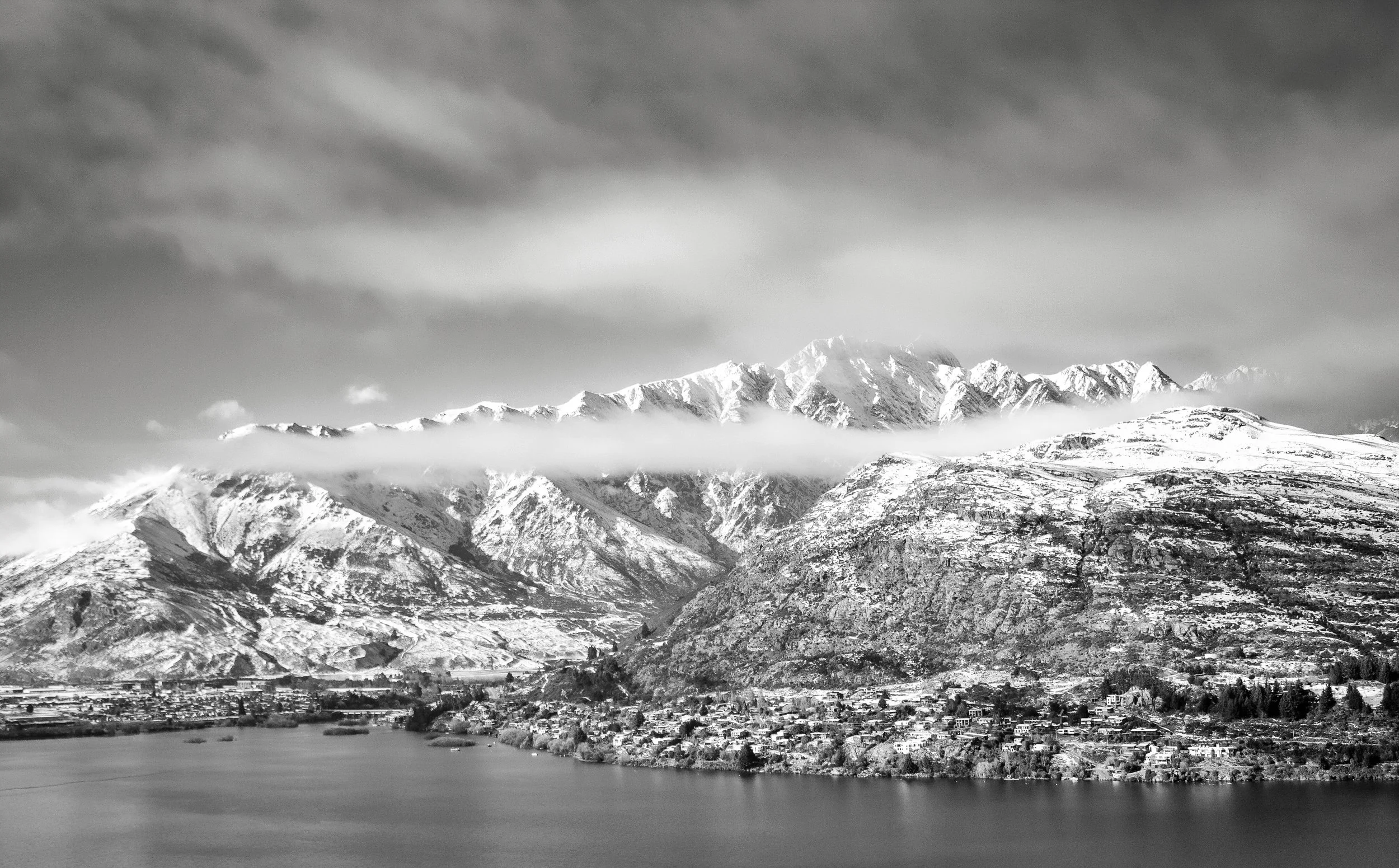 Black-and-white photo of a mountain range with peaks covered in snow, partially shrouded by clouds, with a body of water and a small town in the foreground.