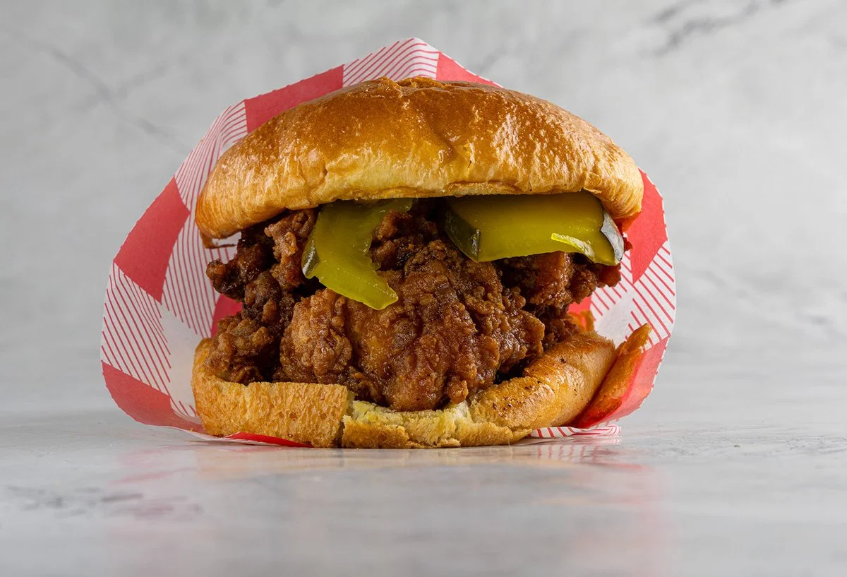 A fried chicken sandwich with pickles in a sandwich bun, wrapped partially in red and white checkered paper, on a light-colored surface.