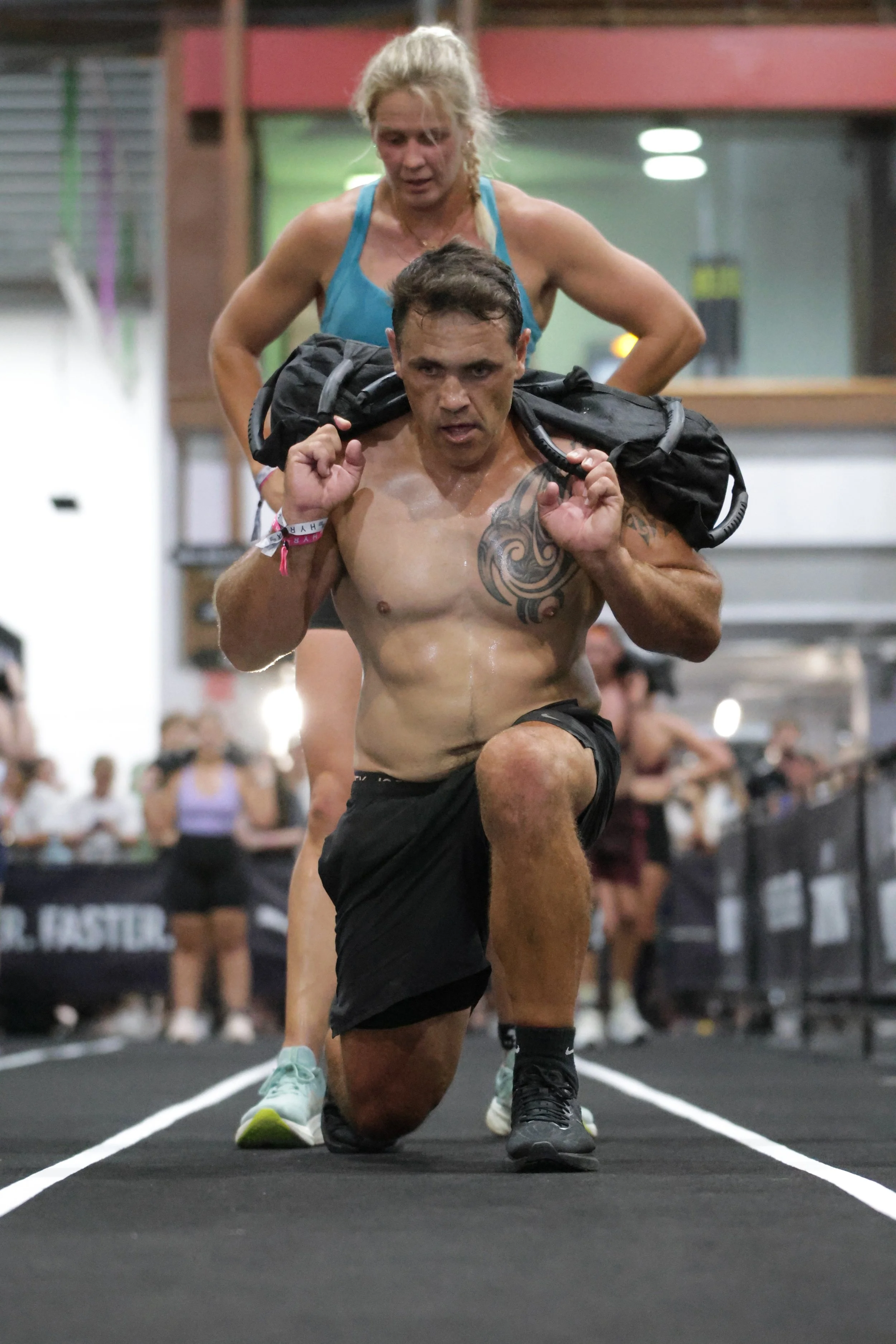 A muscular man kneels on one knee in a gym, carrying a black backpack on his shoulders, with a concentration expression. A blonde woman in a blue sports bra stands behind him, looking down at him. The background has several people and gym equipment.