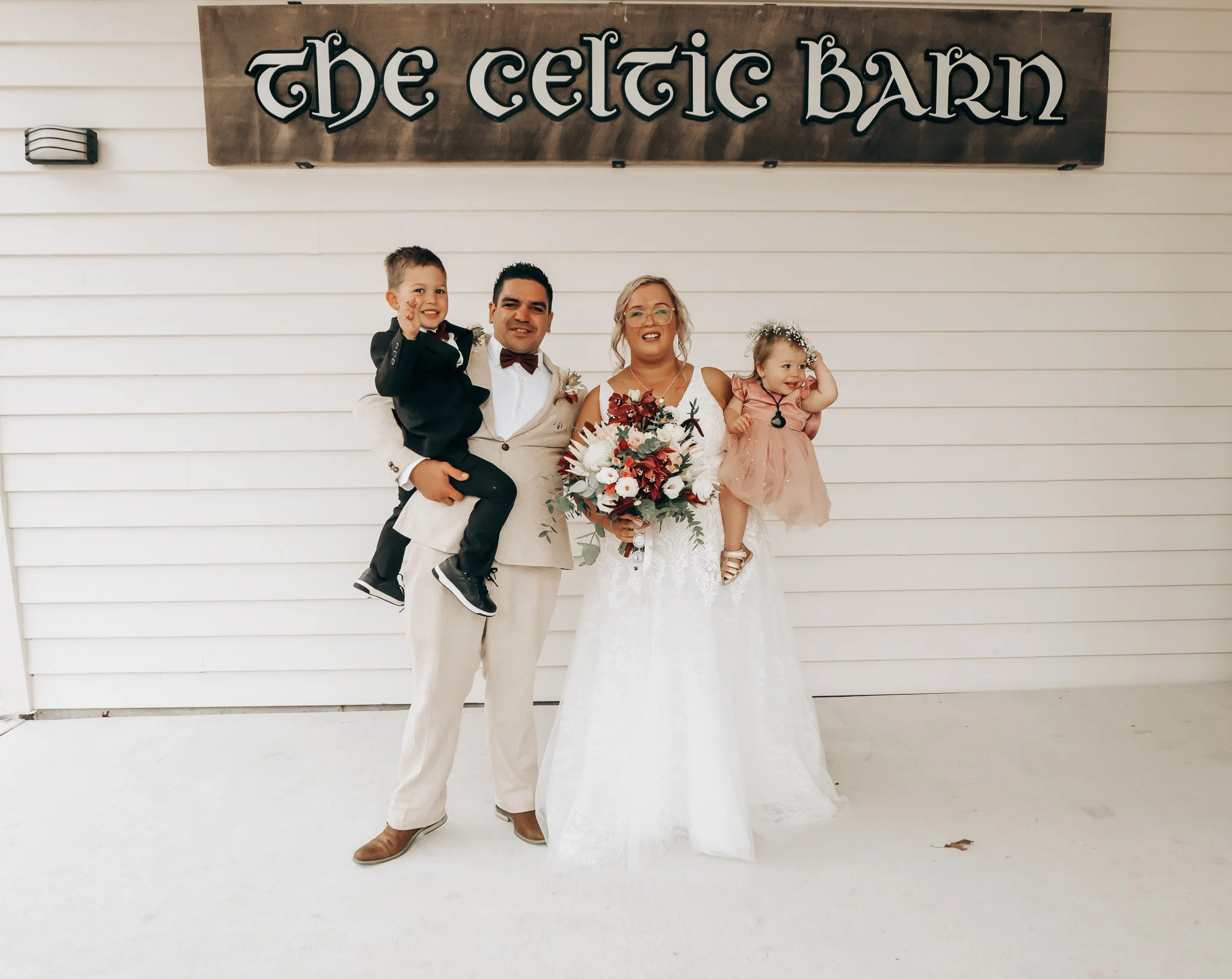 A newlywed couple with two young children standing in front of a sign that says "The Celtic Barn". The man is holding a young boy dressed in a tuxedo, and the woman is holding a bouquet of flowers with a young girl in a light pink dress standing next