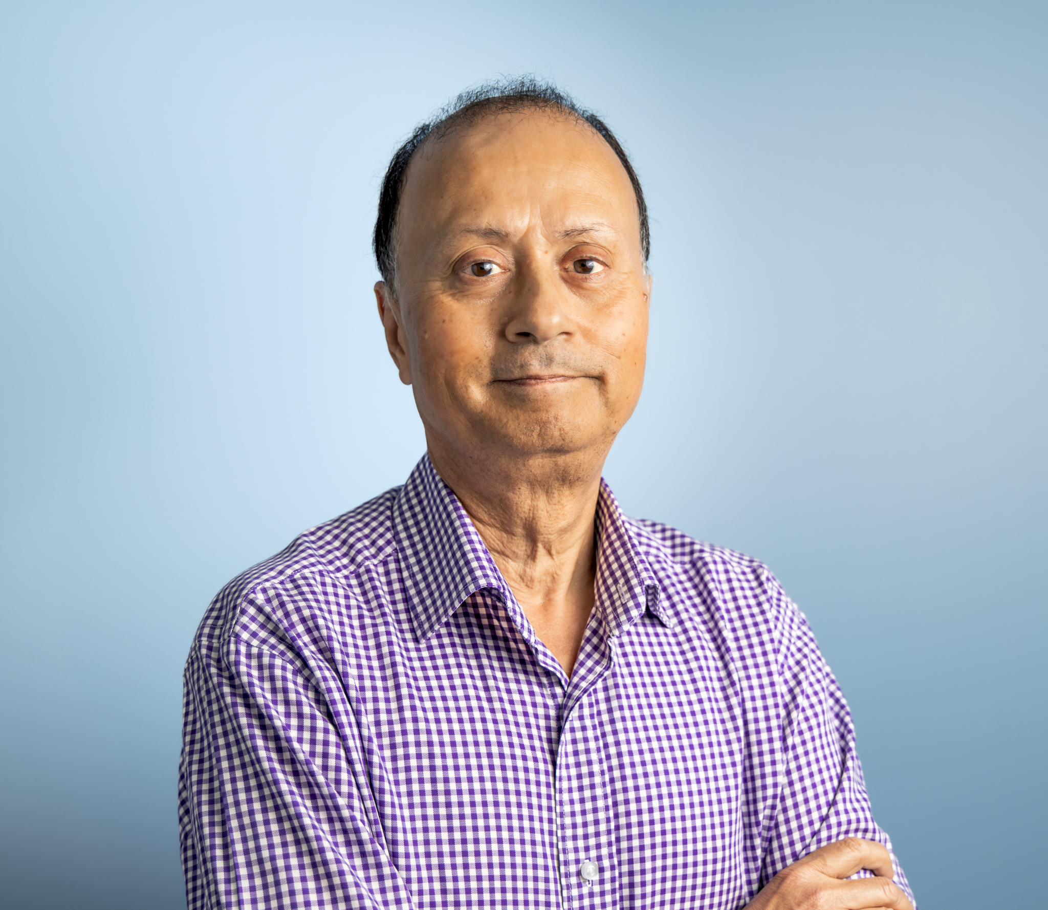 Close-up of a middle-aged man with a receding hairline, wearing a purple and white checkered shirt, standing against a solid light blue background.