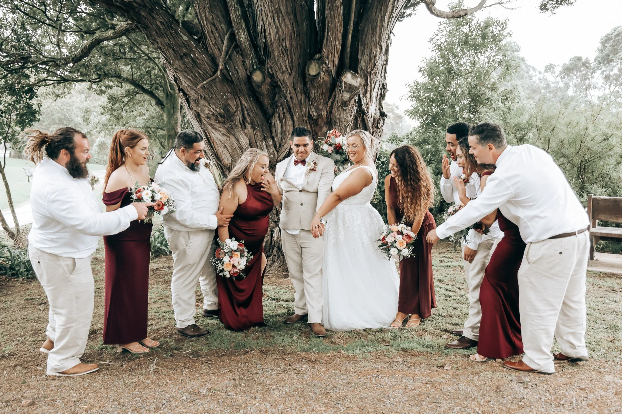 A wedding ceremony with the couple and guests standing outdoors near a large tree, all dressed in formal attire and holding bouquets.