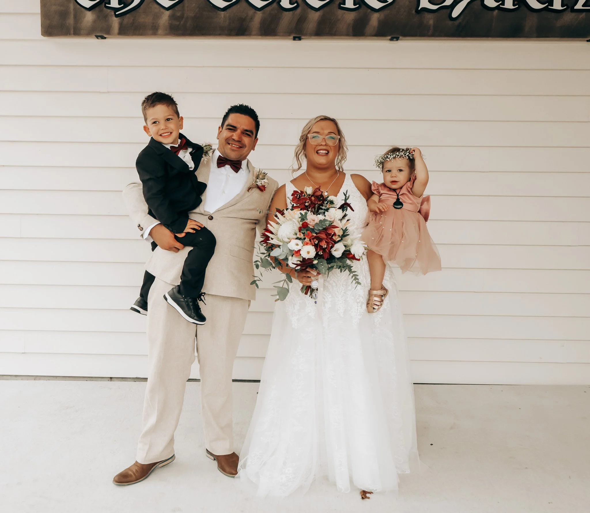 A family of four at a wedding. The father is holding a young boy in a suit, the mother is in a wedding dress holding a bouquet, and a young girl is dressed in a pink dress with a flower crown. They are standing in front of a white wooden wall.