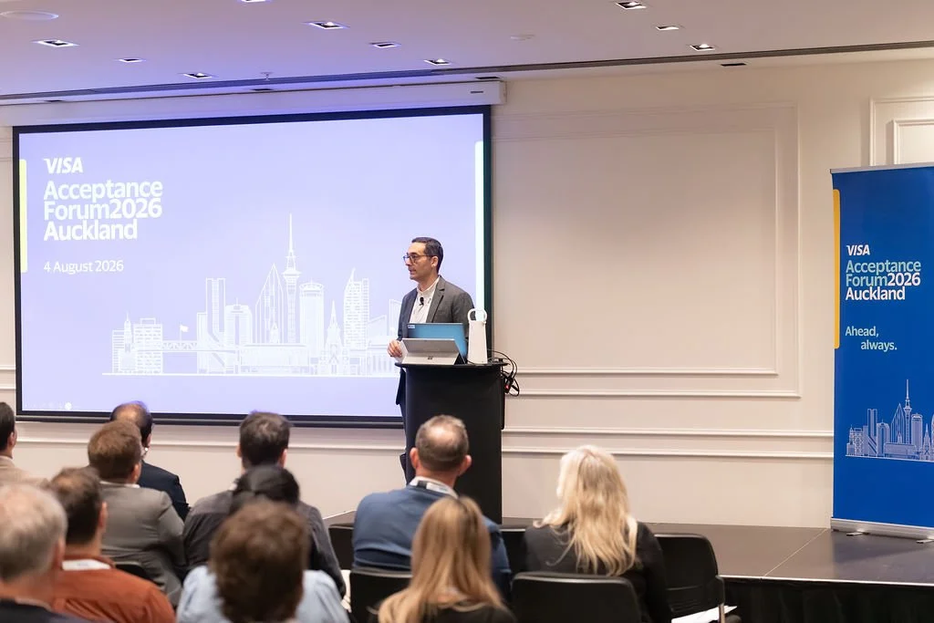 A speaker presenting at the VISA Acceptance Forum 2026 in Auckland, with a slide showing the event name, date, and a city skyline graphic. Audience members are seated and listening.