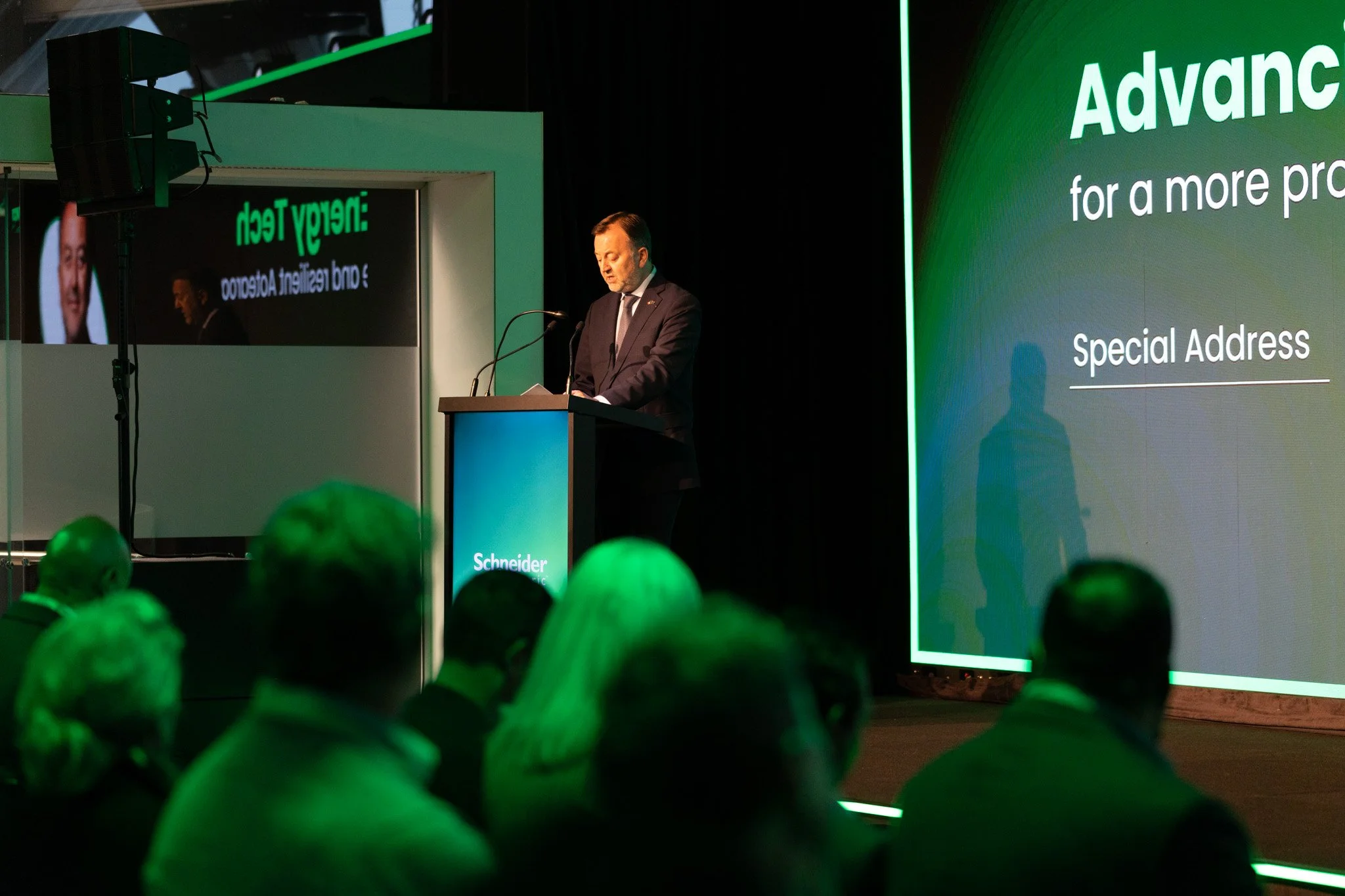 A man in a suit speaking at a podium during a conference with large green presentation screens behind him and an audience visible in the foreground.